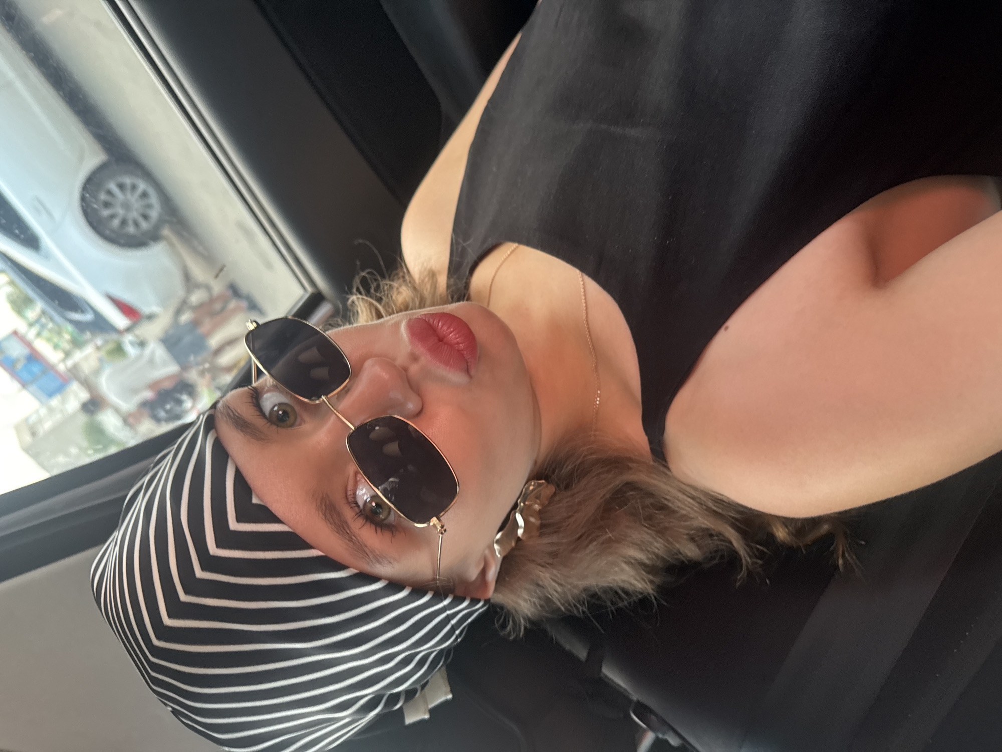 In the city of Chaikovsky, Russia, a woman in a black tank top and a black and white striped headscarf is taking a selfie in what appears to be the backseat of a car. She is wearing gold-rimmed square sunglasses and large gold earrings shaped like flowers. Her lips are pursed, and her right arm is visible as she holds the phone. Through the car window, a white car and pedestrians are visible in the background, suggesting an outdoor urban setting. The lighting is natural, indicating daylight.