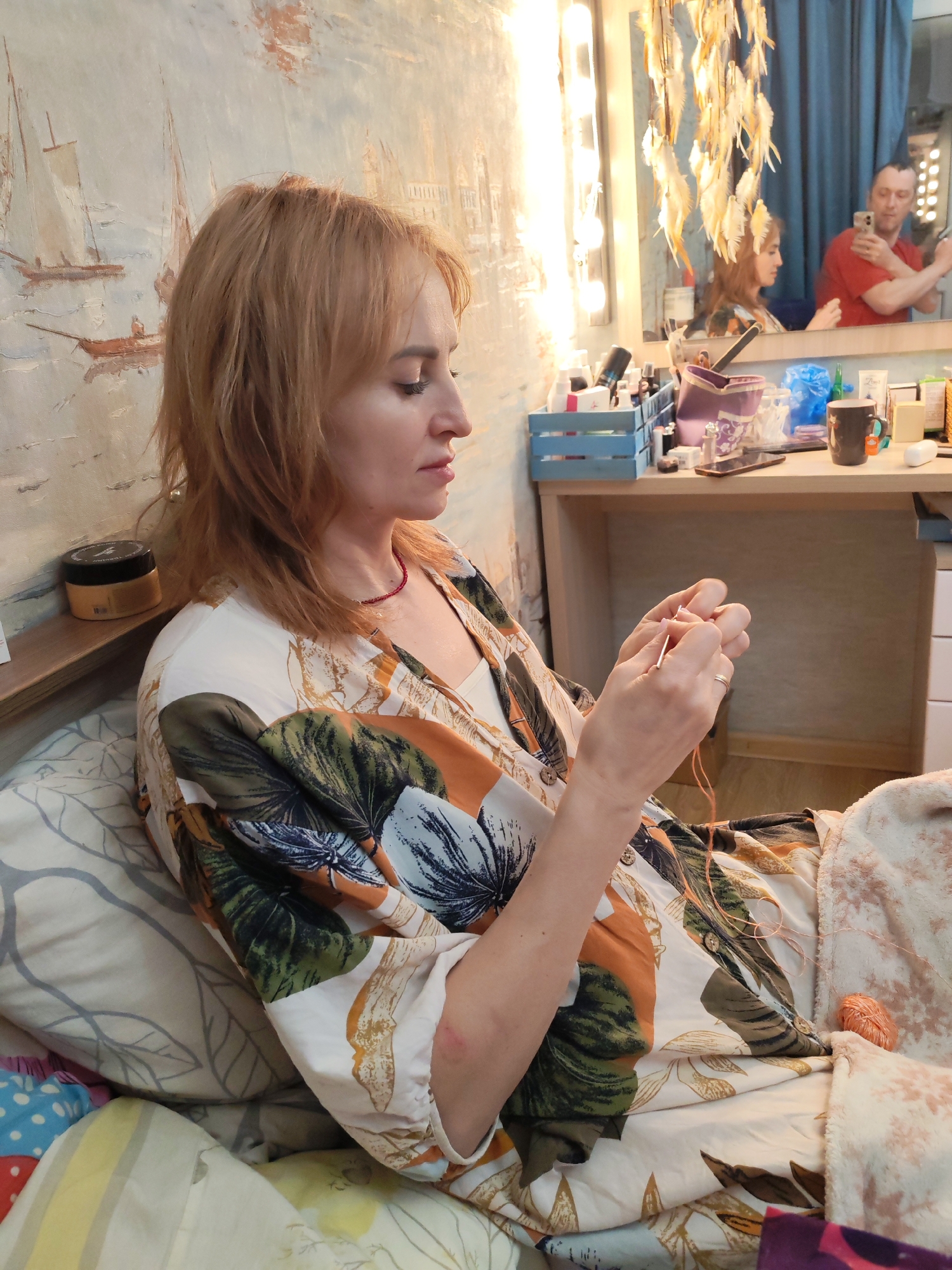 The image captures an indoor scene within the city of Chaikovsky, Russia. A fair-skinned woman with reddish-blonde hair, wearing a loose-fitting white top with large green, brown, and orange leaf patterns, is seated, viewed in profile, looking downwards. She is actively engaged in knitting or crocheting, holding a slender needle and orange yarn with both hands. A red beaded necklace is visible around her neck, and a plain gold ring is on her left ring finger. A faint reddish mark is present on her right elbow.
She is situated on patterned bedding, including a pillow with grey leaf motifs. To her immediate left, a narrow wooden shelf holds a dark container with a light-colored lid. The wall behind her features a light-toned mural or wallpaper depicting sailing boats on water and distant structures.
To the right, a vanity or dressing table is visible, topped with numerous personal care items, cosmetics in jars and bottles, a purple fabric bag, blue storage crates, a black mug, and a dark smartphone. Above the table, a rectangular mirror is mounted, framed by vertical strips of bare light bulbs. Hanging in front of the mirror is a decoration resembling a dreamcatcher, made of light-colored feathers. The mirror's reflection shows a blue curtain in the background, the woman's profile, and a fair-skinned man with short hair wearing a red t-shirt. The man is holding a silver-colored smartphone up, appearing to be taking a photo or video of the scene. A loose ball of orange yarn rests on the bedding to the woman's right.