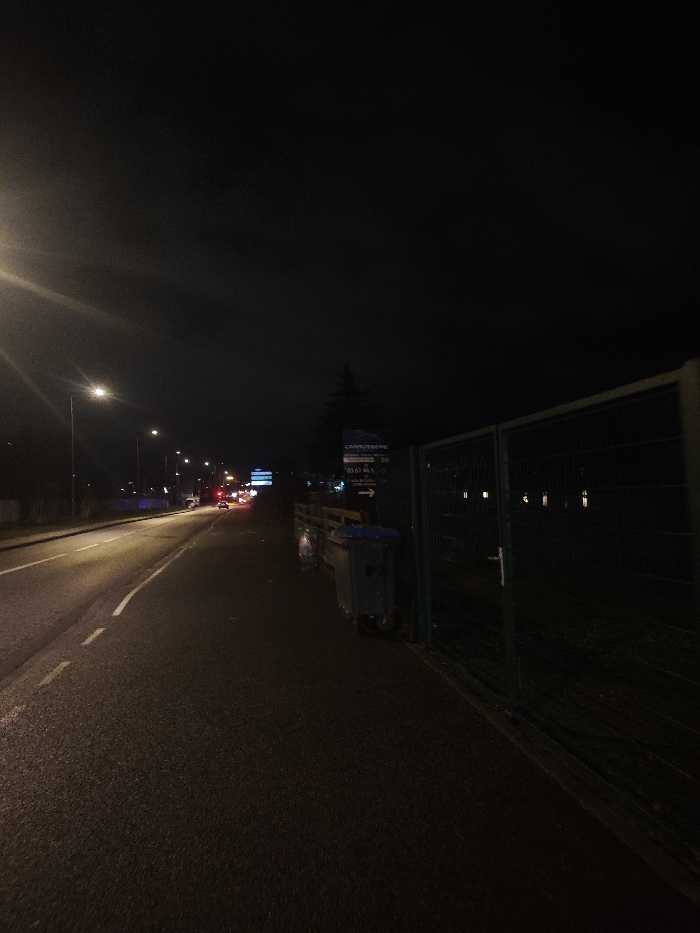 A nighttime street scene shows a dark road illuminated by streetlights leading towards distant car lights. A sidewalk runs along the right, bordered by a green chain-link fence. Adjacent to the fence are overflowing trash bins and a signpost with business information. The sky is dark and overcast, adding to the somber, urban atmosphere.