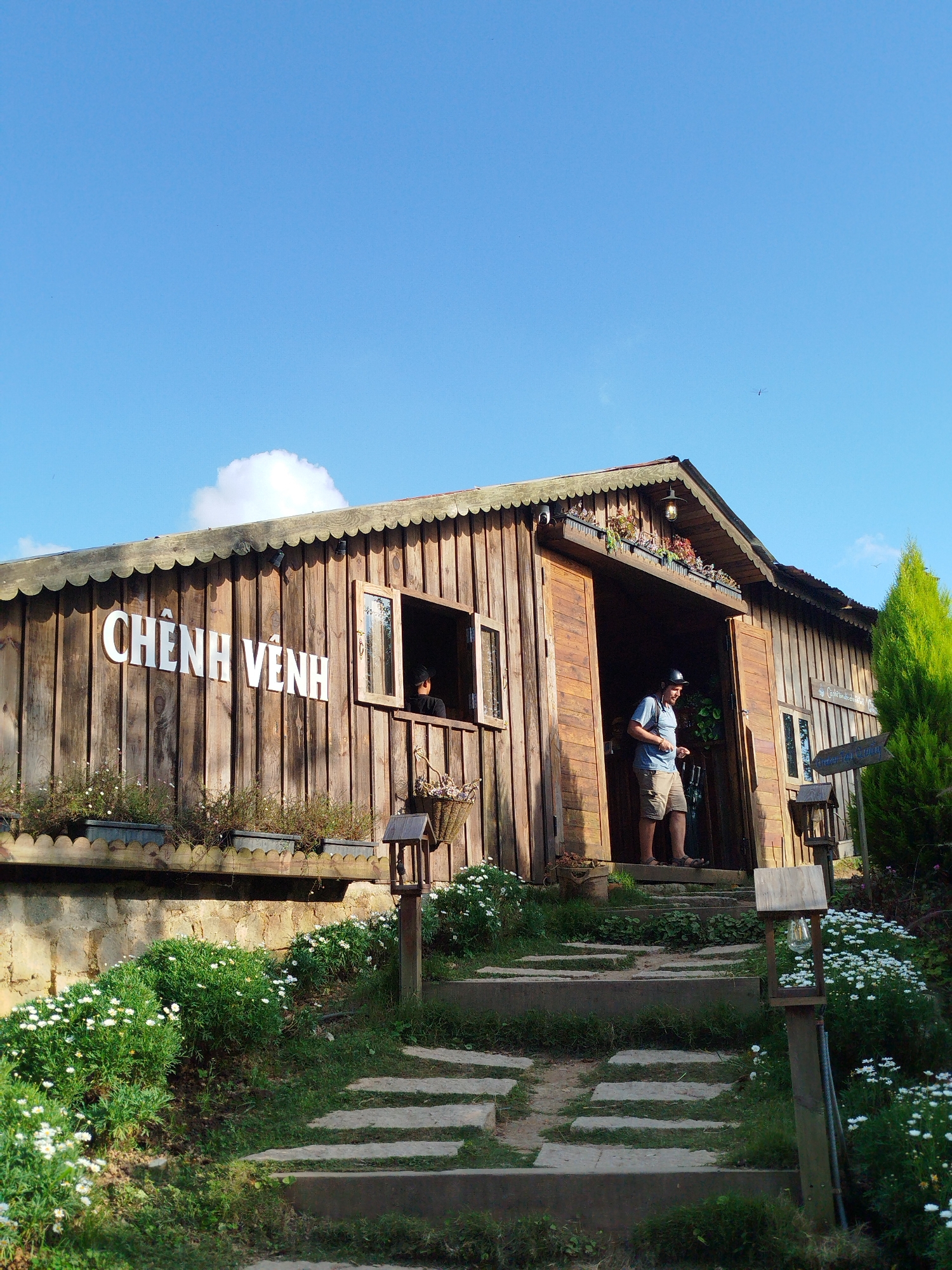 The image depicts a wooden building with the Vietnamese signage "CHÊNH VÊNH" on its facade, situated outdoors. A man wearing a blue t-shirt and shorts stands in the open doorway of the building, holding a mobile phone. In front of the building, a stone pathway leads upwards, flanked by lush green grass and small white flowers. The building has windows, one of which shows a person inside. A tall green coniferous tree stands to the right of the building. The sky above is a clear, bright blue with a single white cloud. The overall scene suggests a rustic, possibly tourist-oriented location. Given the signage and the presence of flowers, the location is likely a cafe or shop in the Lâm Viên area of Đà Lạt, Vietnam.