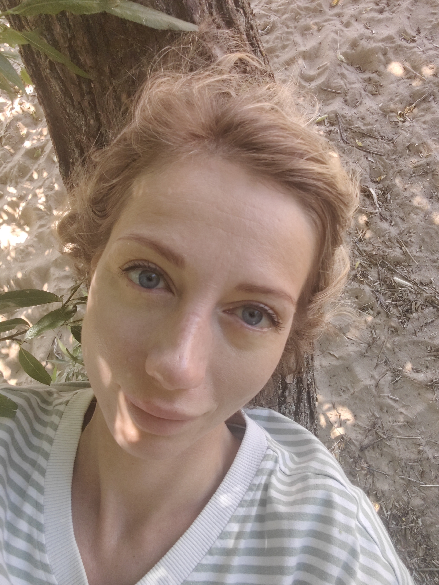 A close-up, selfie-style photograph shows a fair-skinned woman with light blonde, curly hair. She is wearing a light green and white striped long-sleeved shirt. Her face is turned slightly towards the camera, and she has blue eyes, light eyebrows, and her lips are closed. The background consists of a tree trunk to her left, with green leaves visible in the upper left corner. The ground behind her appears to be sandy with small twigs and dry leaves scattered about. The lighting suggests an outdoor setting, possibly in a park or natural area. No specific city location is identifiable from the visual information provided.