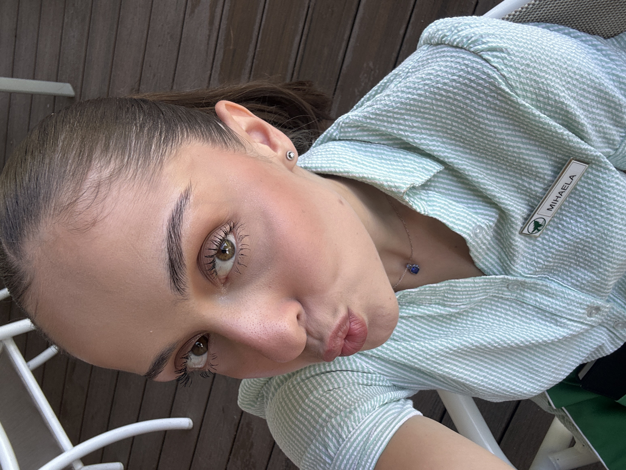 A young woman with her hair pulled back in a ponytail, wearing a mint green and white striped collared shirt and a name tag that reads "MIHAELA," leans in towards the camera, puckering her lips. She has on subtle makeup, including mascara, eyeshadow, and blush, and is wearing a delicate necklace with a blue pendant and small stud earrings. The background suggests an outdoor setting with a wooden deck and white outdoor furniture. The overall impression is a casual, selfie-style photograph.