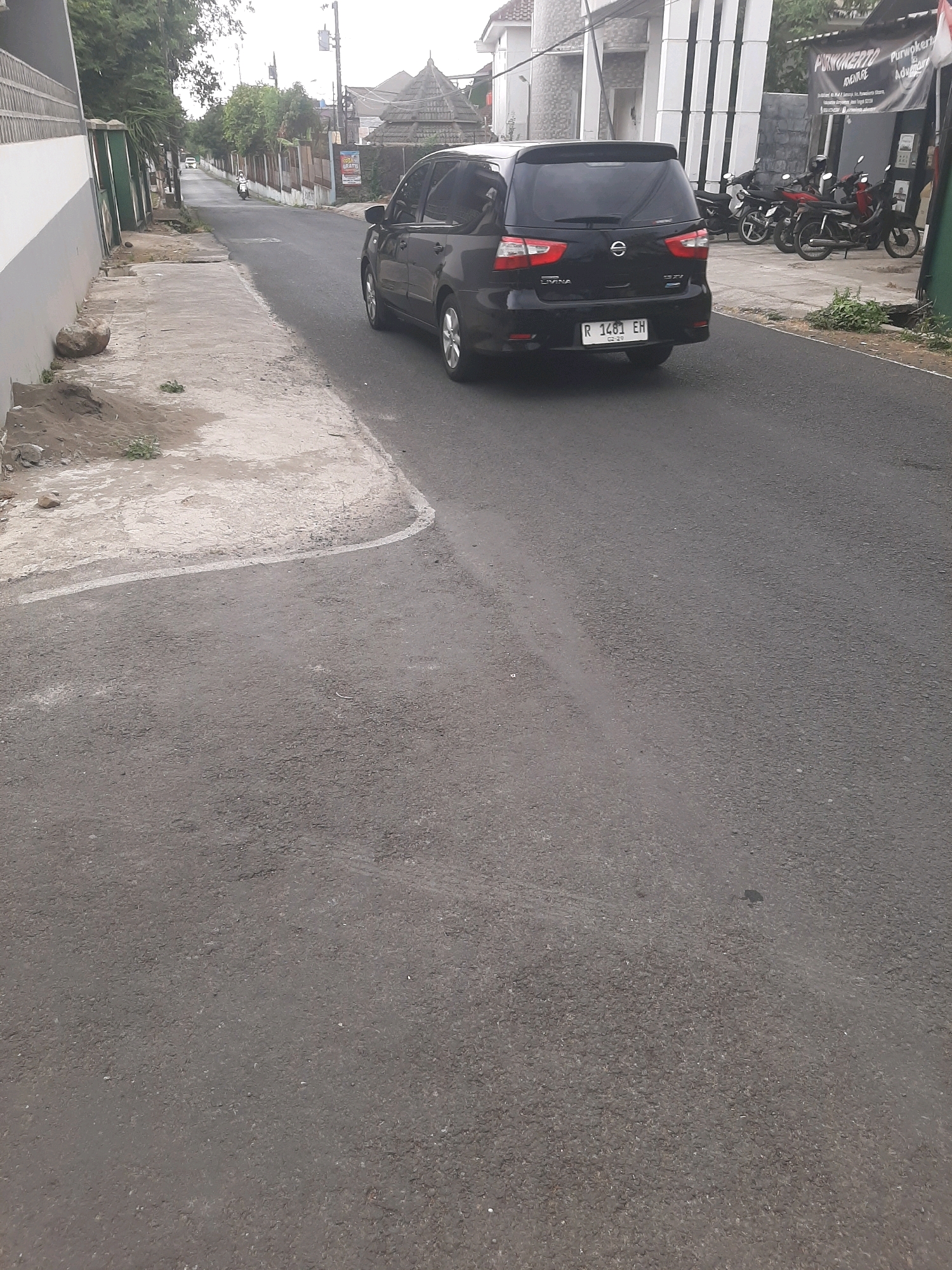 A black Nissan Grand Livina is parked on the right side of an asphalt road in Sumampir, Indonesia. The car's license plate reads "R 1483 EH". To the left of the road, a concrete sidewalk is visible, bordering a low wall. In the background, several motorcycles are parked in front of a building with "PURWOKERTO ADVENTURE" written on a banner. The sky is overcast, and trees line the street. The overall scene depicts a typical urban street in Indonesia.
