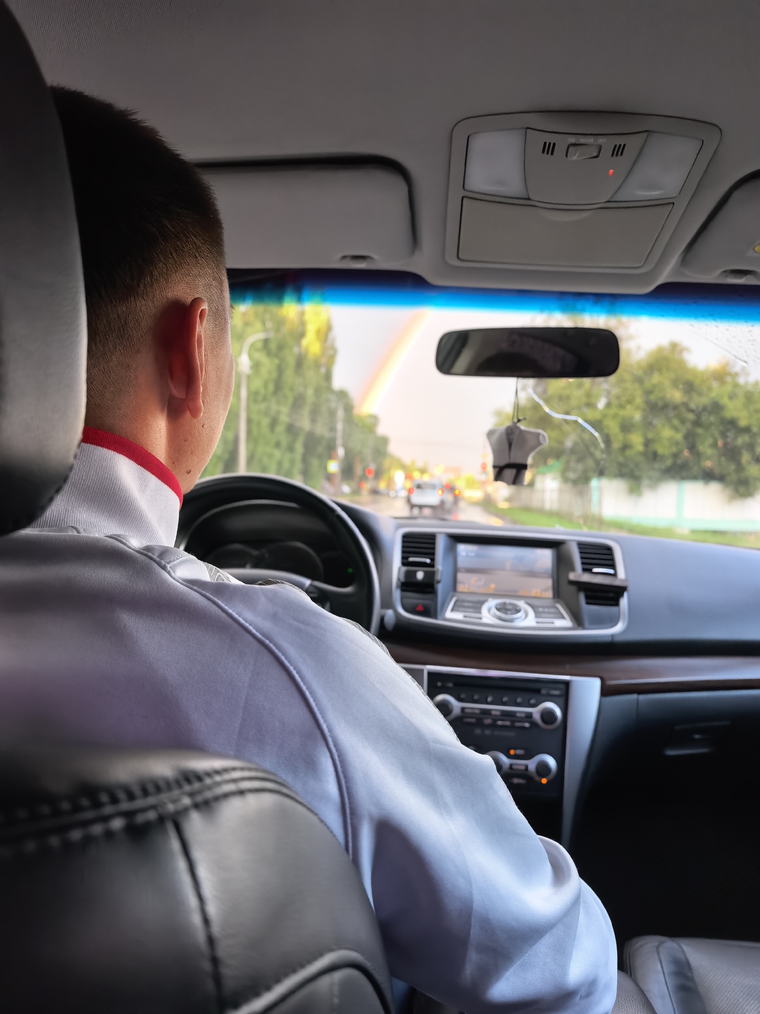 The image shows a man driving a car, viewed from behind. He is wearing a white shirt with a red trim around the collar. The car's interior is visible, including the dashboard, steering wheel, and a navigation screen. Outside the car, a rainbow is visible in the sky, along with trees and streetlights, suggesting it is a residential area or a road in a city. The license plate of the car is not visible, so its specific location cannot be identified, however the presence of a rainbow suggests recent rainfall.