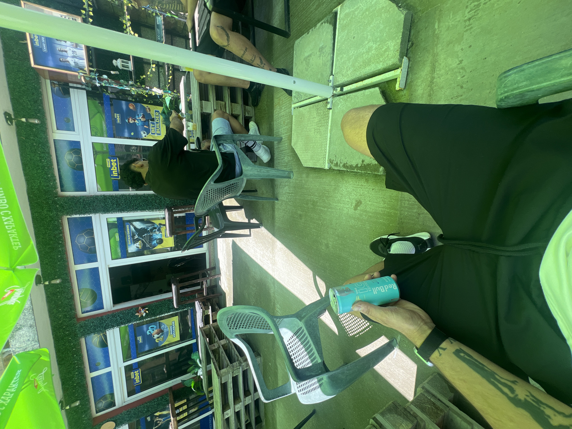 The scene depicts an outdoor patio area in Касапите, Bulgaria, under a green canopy that casts a green tint over the concrete floor. In the foreground, a person is seated, wearing black shorts, black shoes, and a smartwatch on their left arm, which bears a tattoo. This individual holds an open light blue "Red Bull THE ICED EDITION" can. To the left, a second person is seated in a green plastic chair, facing away, wearing a black t-shirt, light blue shorts, white socks, and white sneakers. They are positioned at a low table constructed from wooden pallets, on which a glass containing a yellow liquid is visible. To the right, a third person's right leg, also tattooed, is partially visible, clad in black shorts and black shoes, seated at the same pallet table. A white pole, likely supporting the canopy, stands anchored by four concrete slabs on the ground. The background features a building with windows and an entrance, adorned with artificial green foliage. Advertisements for "inbet," a betting company, are prominently displayed on posters within and around the windows, featuring sports themes such as basketball and soccer, with phrases like "BET BUILDER" and "ЕКСКЛУЗИВНО" (exclusive) in Cyrillic. A white lantern and some draped artificial vines are visible on a shelf unit behind the seated individuals. Sunlight filters through the canopy, creating distinct shadows on the ground.