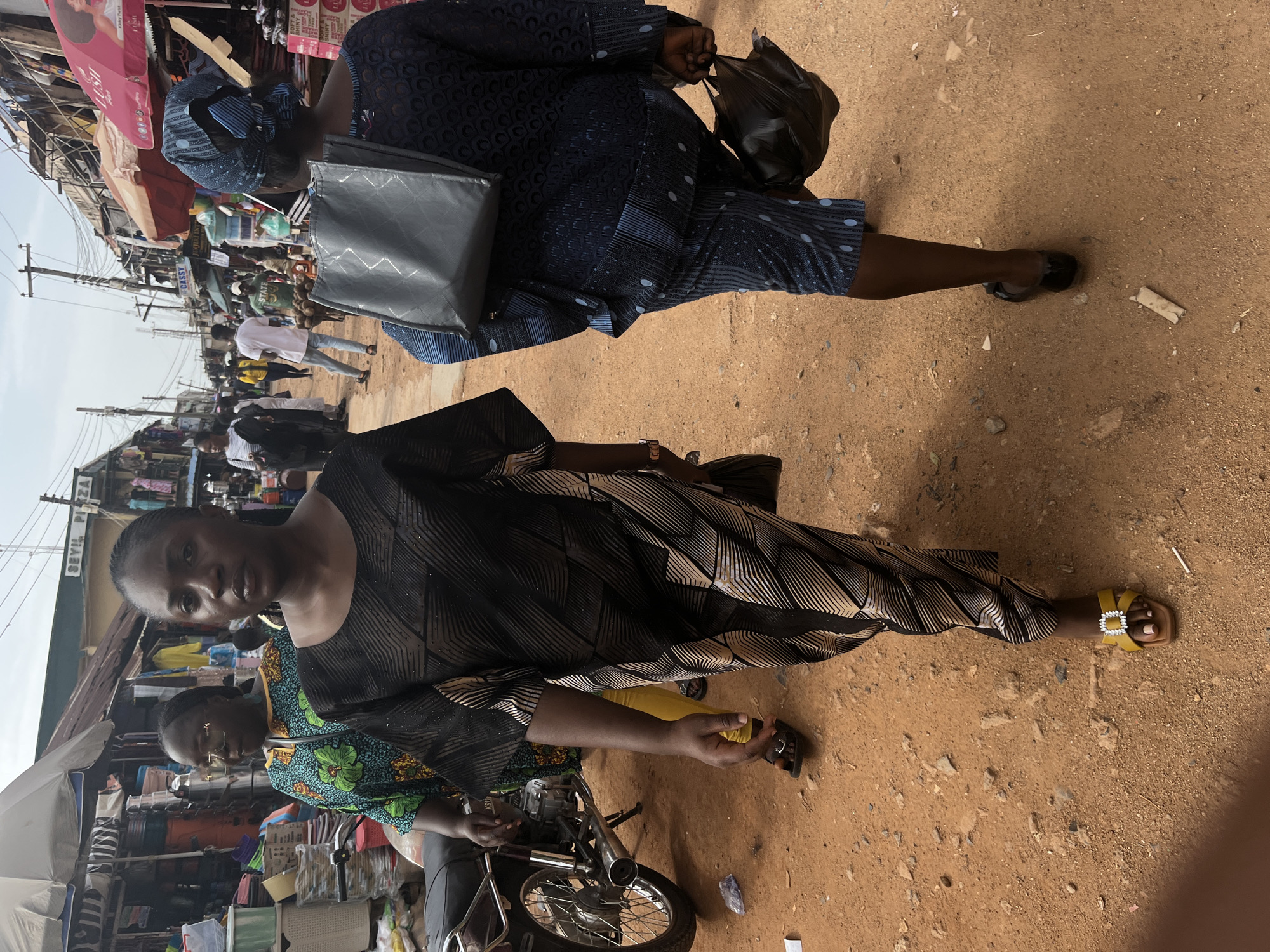 This image captures a bustling street scene in Jos, Nigeria. The primary subjects are two women, one in the foreground and another further back. The woman in the foreground is wearing a black and gold patterned dress and yellow sandals. She is looking towards the camera. Behind her, a second woman is dressed in a navy blue patterned outfit and a matching headwrap. She is carrying a black bag in her right hand and a large grey bag on her left side.
The scene is a busy marketplace with stalls and various goods visible. In the background, other people can be seen walking and going about their business. A motorcycle is parked in the lower right portion of the image, suggesting transportation is common in this area. The ground is unpaved, indicating a typical street environment in this region. The lighting suggests it is daytime, with bright sunlight casting shadows.
There is some visible text on signs in the background, including "STATE" on a green shirt worn by someone in the midground, and partially obscured text that appears to be part of a shop name. The overall atmosphere appears to be one of lively commerce and daily life in a Nigerian urban setting.