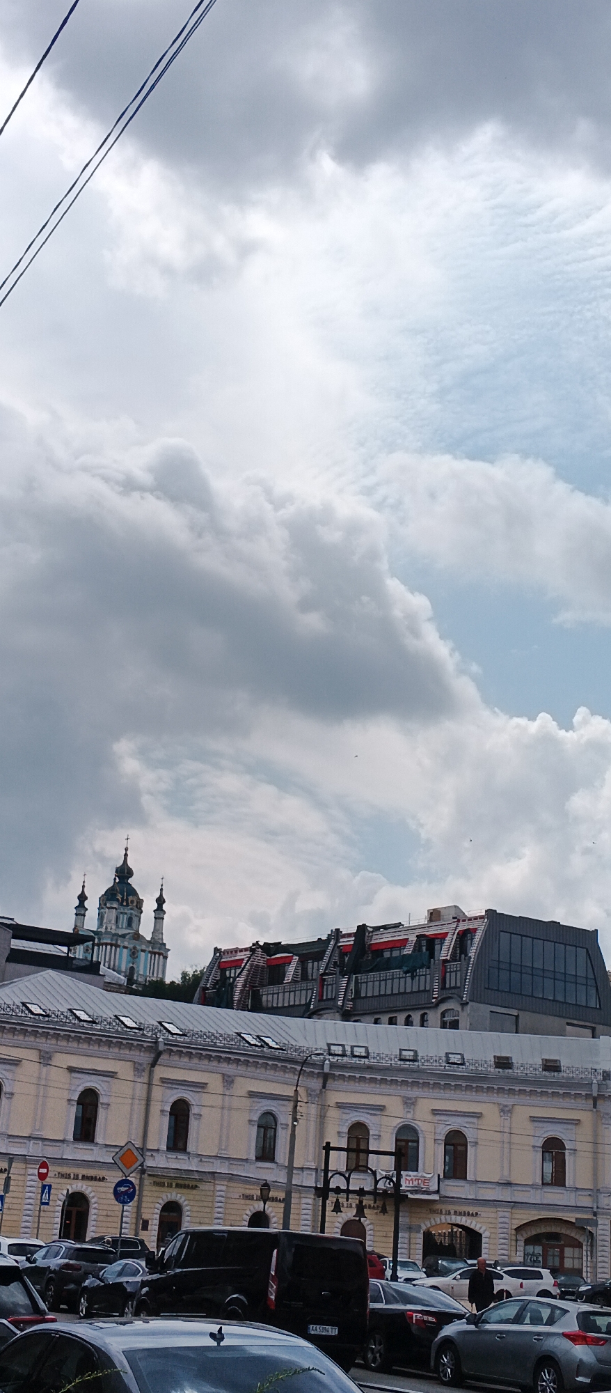 The image shows a street scene in Kyiv, Ukraine, with a view of St. Andrew's Church in the background, a prominent blue and white domed orthodox church. In the foreground, there is a row of buildings with light yellow facades and arched windows. A modern building with a glass facade and a red-accented roof is visible behind the yellow buildings. Several cars are parked on the street, and a few people are visible walking. Overhead, power lines cross the sky, which is filled with dramatic, cloudy formations. The scene appears to be captured during the daytime.