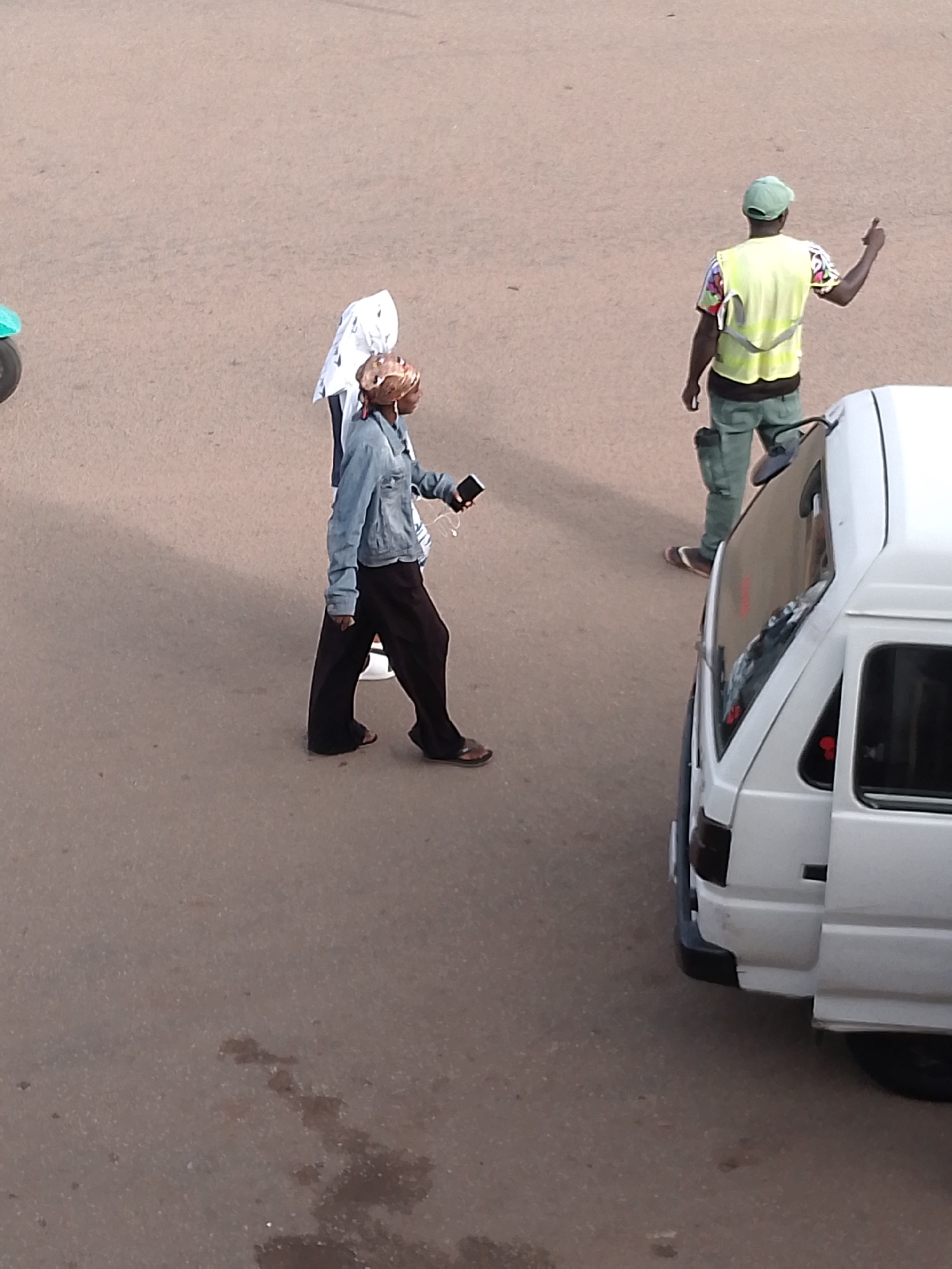 In Bukuru, Nigeria, a medium shot captures a street scene from an elevated perspective. In the foreground, the rear of a white minivan is visible. To the left of the minivan, a woman walks away from the camera, holding a smartphone in her right hand and wearing earbuds. She is dressed in a light blue denim jacket, dark wide-leg pants, and sandals, with a patterned headscarf. Behind her, another person, partially obscured, appears to be walking in the same direction, wearing a white garment. Further in the background, to the right, a man wearing a green cap, a light yellow reflective vest over a brightly patterned shirt, and green pants stands facing away from the camera and gestures with his right hand. The ground is paved with a reddish-brown surface, indicative of a dirt or asphalt road, with some scattered debris and a wet stain visible. The lighting suggests it is daytime.