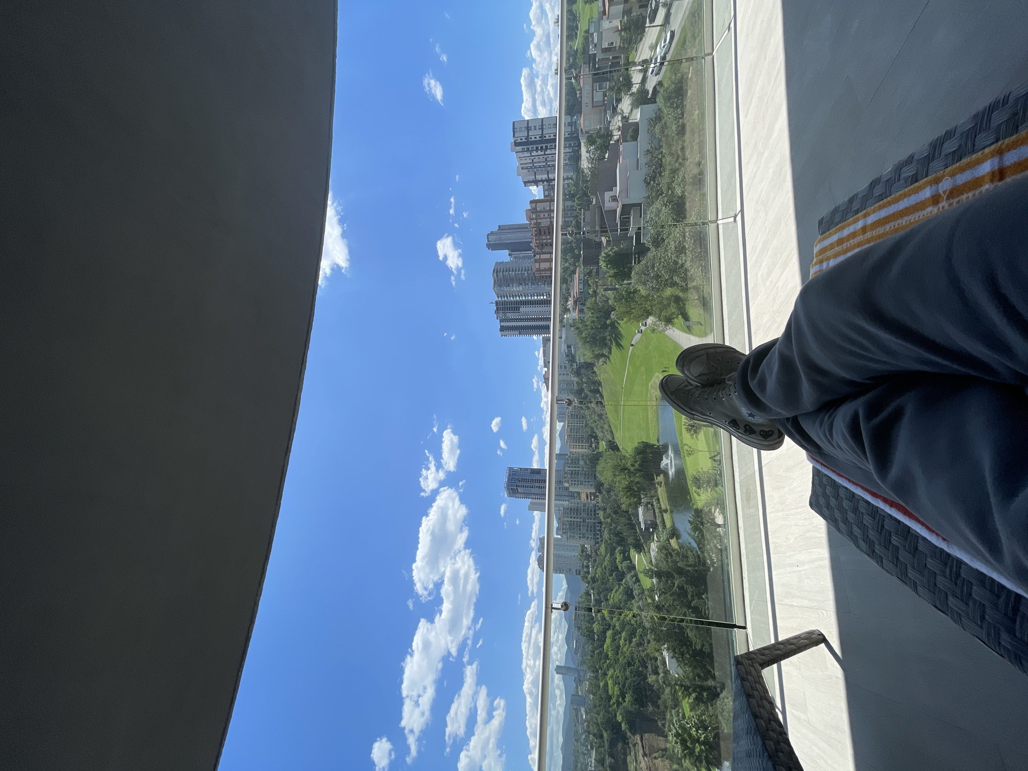 Here is a forensic description of the image:
The image is a medium shot taken from a high vantage point, likely a balcony, in Huixquilucan de Degollado, Mexico. The camera is positioned at an angle, capturing a partial view of a person resting on a wicker chaise lounge. The person's legs, clad in gray sweatpants and gray sneakers, are crossed. A striped towel lies on the chaise lounge.
Beyond a glass balcony railing, the view extends to a cityscape. Buildings of varying heights are visible, interspersed with greenery. The sky is blue with scattered white clouds. A body of water with a fountain is visible within the green landscape. The environment suggests an urban setting with recreational spaces.