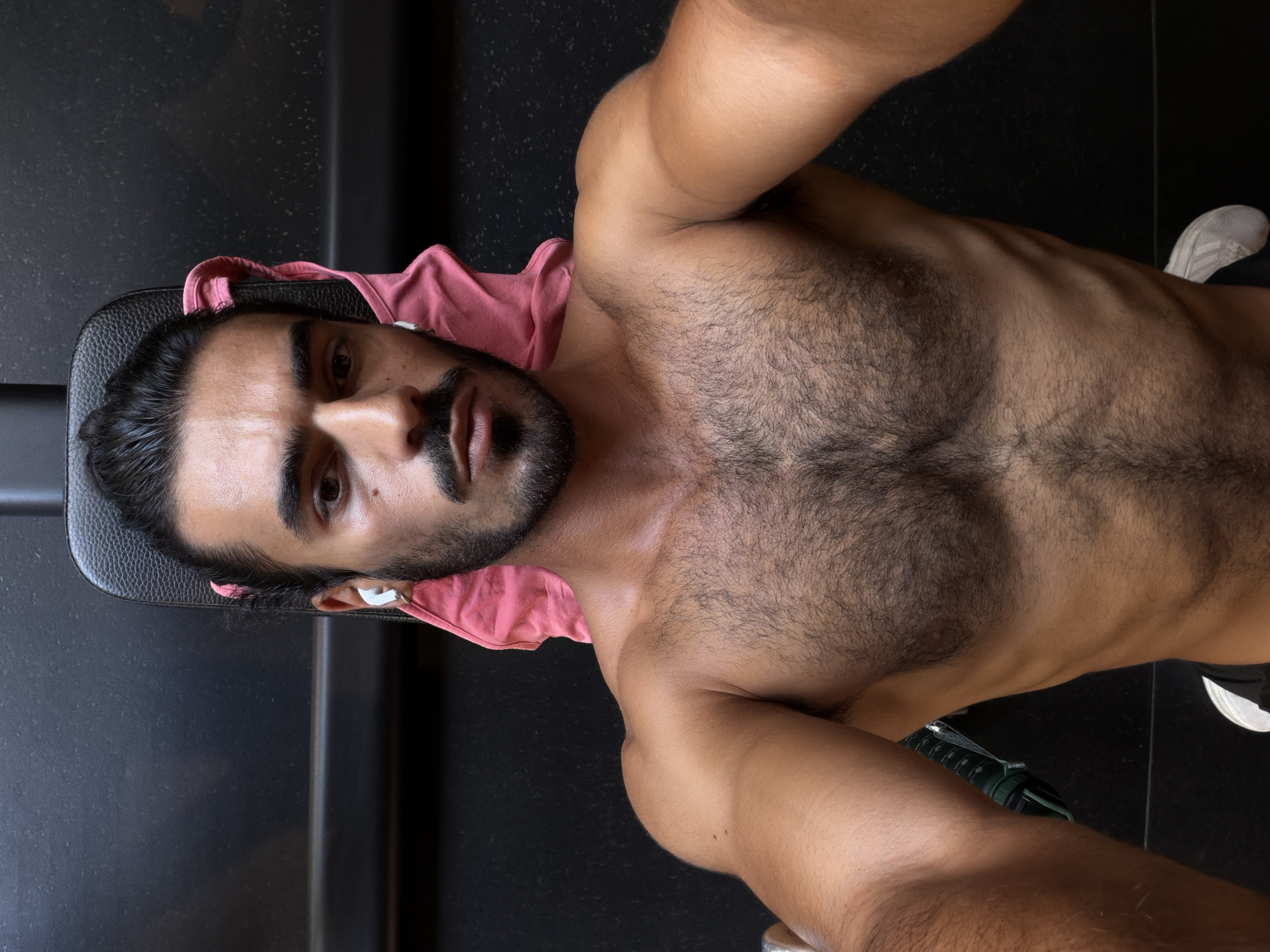A shirtless man with dark hair, tied in a bun, and a beard is sitting on a black gym bench. He is wearing white earbuds and has a pink towel draped over the back of the bench behind him. The background is dark and appears to be a gym environment. The man is looking directly at the camera.