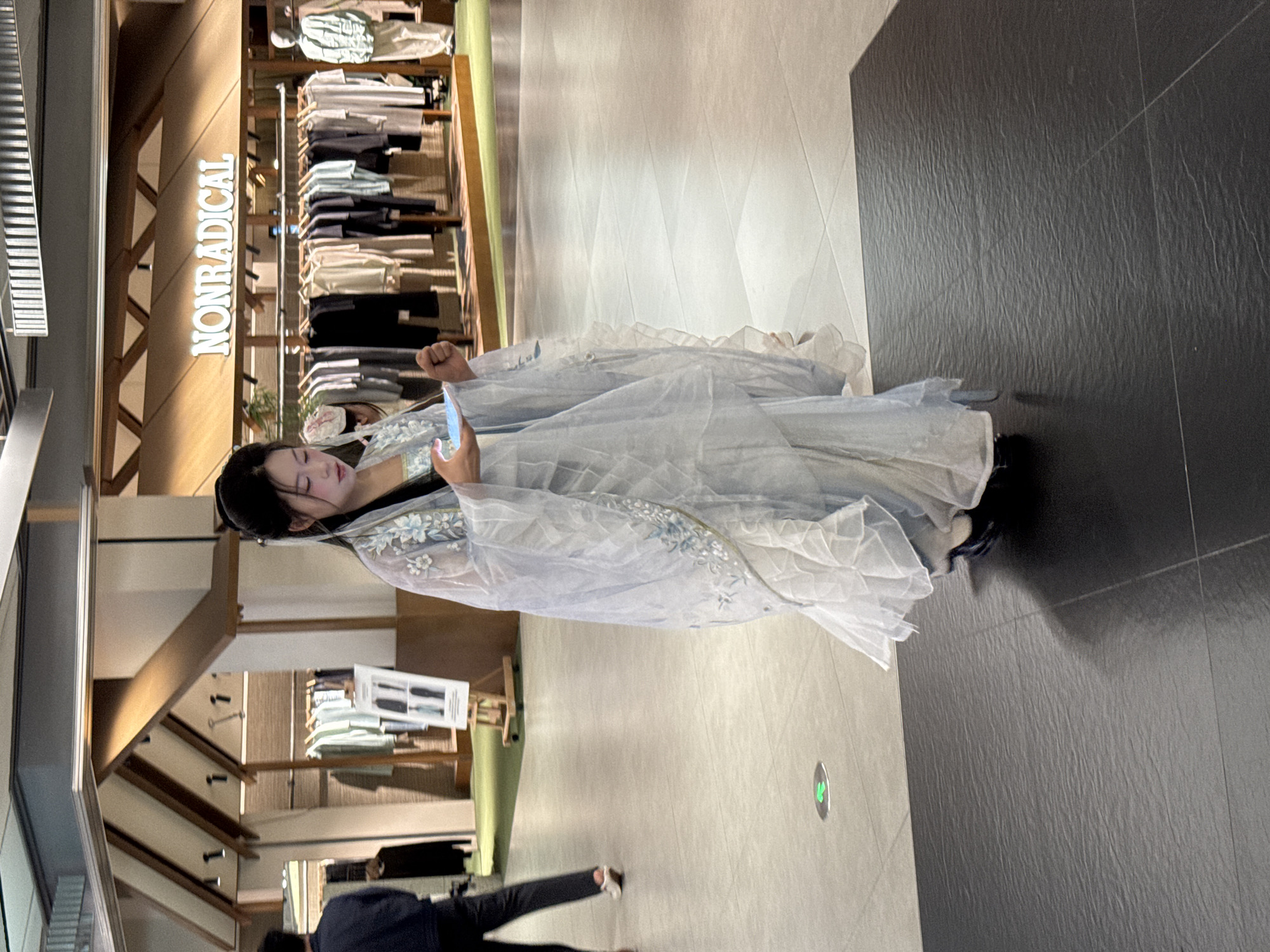 In a brightly lit commercial space, a woman dressed in a flowing, light blue and white hanfu stands holding a smartphone, seemingly interacting with it. The location is likely a shopping mall given the presence of clothing racks and a store sign that reads "NONRADICAL." A person is walking by in the lower part of the frame. The floor is light-colored tile near the woman and transitioning to dark tile on the right side. The overall environment suggests an interior setting within a modern commercial area, possibly in Jinjiang District, China.