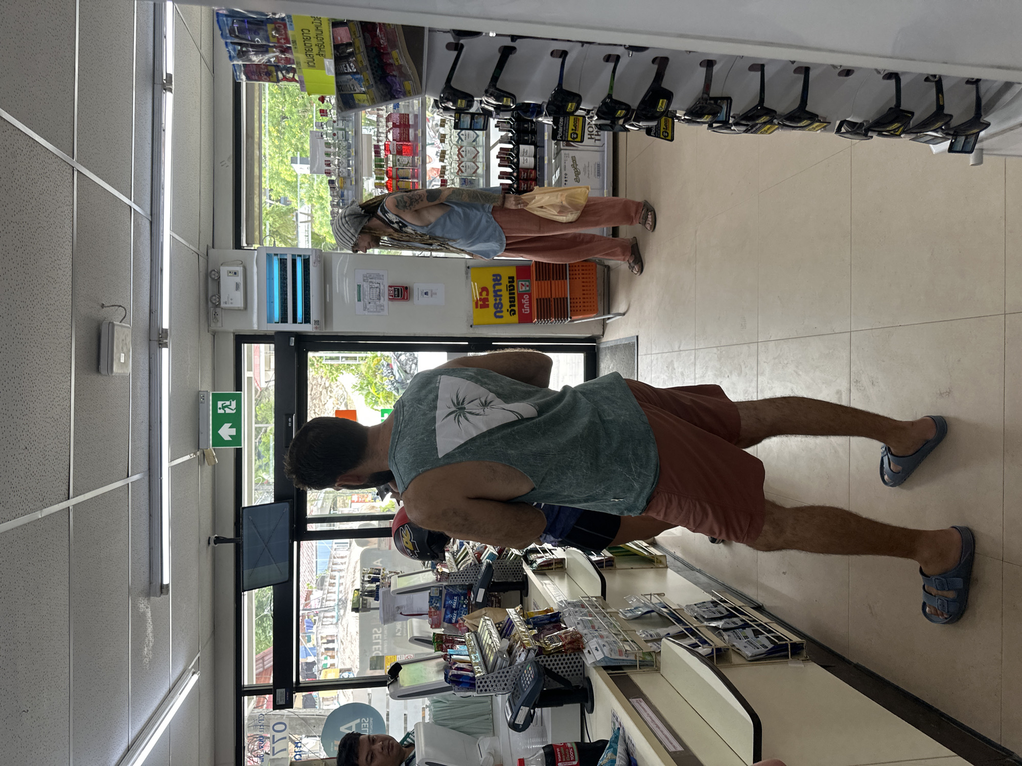 In a convenience store in Tambon Koh Phangan, Thailand, three individuals are observed. In the foreground, a man with dark hair and a beard stands with his back to the camera, wearing a green sleeveless t-shirt with a palm tree design and red shorts. He is barefoot. To his right, a person with dreadlocks, wearing a light blue tank top and tan pants, is looking towards the left. They are holding a light-colored plastic bag in their right hand and wearing sandals. Further left, a man with dark hair and a light green shirt is partially visible, appearing to be an employee behind a counter. The store interior features a tiled floor and ceiling with fluorescent lighting. Shelves stocked with various products, including drinks, snacks, and sunglasses, are visible. A digital display screen and point-of-sale terminals are on the counter. An exit sign with an arrow and a running figure is illuminated above a doorway in the background. A blue insect zapper is mounted on the wall to the right of the doorway.