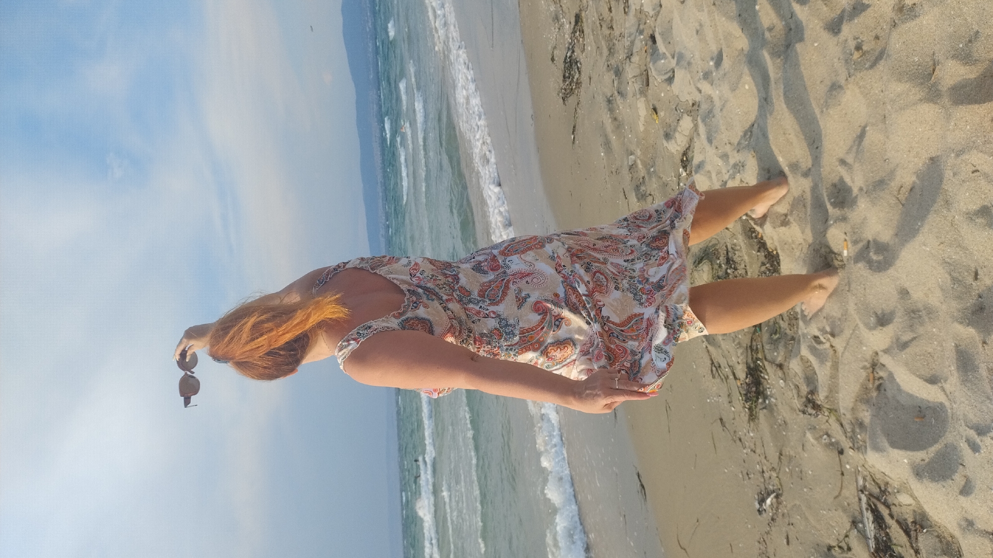A woman with long, auburn hair stands on a sandy beach in Neoi Epivates, Greece. She is wearing a patterned sundress and holding a pair of sunglasses in her right hand. Her left hand is resting on her hip. The woman is facing away from the camera, looking out at the sea. The waves are gentle, and the sky is clear and blue. The sun appears to be high in the sky, suggesting it is midday. The sand is light brown and appears to be mixed with some dried seaweed. The woman's bare feet are visible in the sand. The overall mood of the image is peaceful and relaxed.