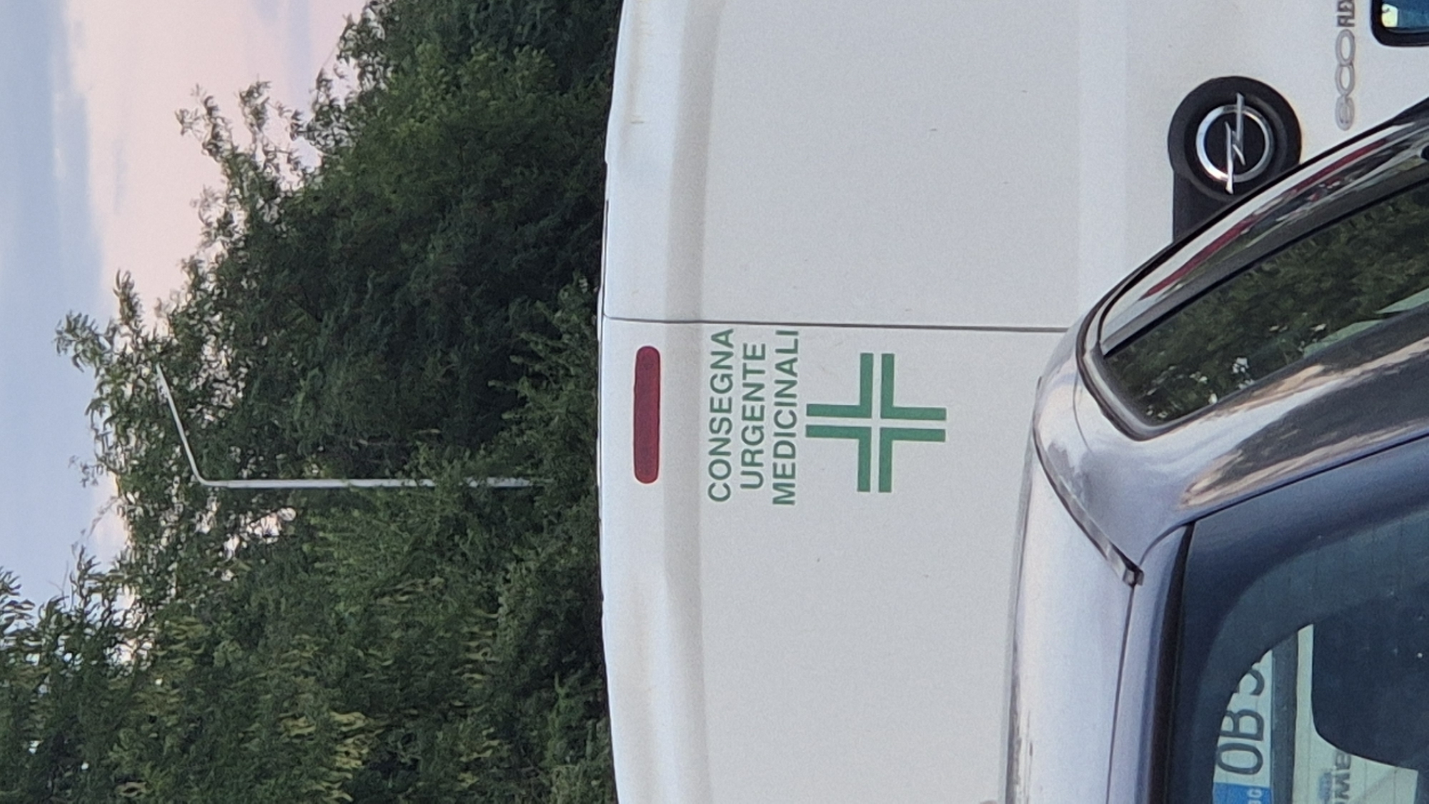 In an urban outdoor setting, a white van is parked behind a dark-colored car. The van has a green cross emblem and text that reads "CONSEGNA URGENTE MEDICINALI" in green. The dark car has some text on its rear window, including "OBS". The scene is captured at eye level.