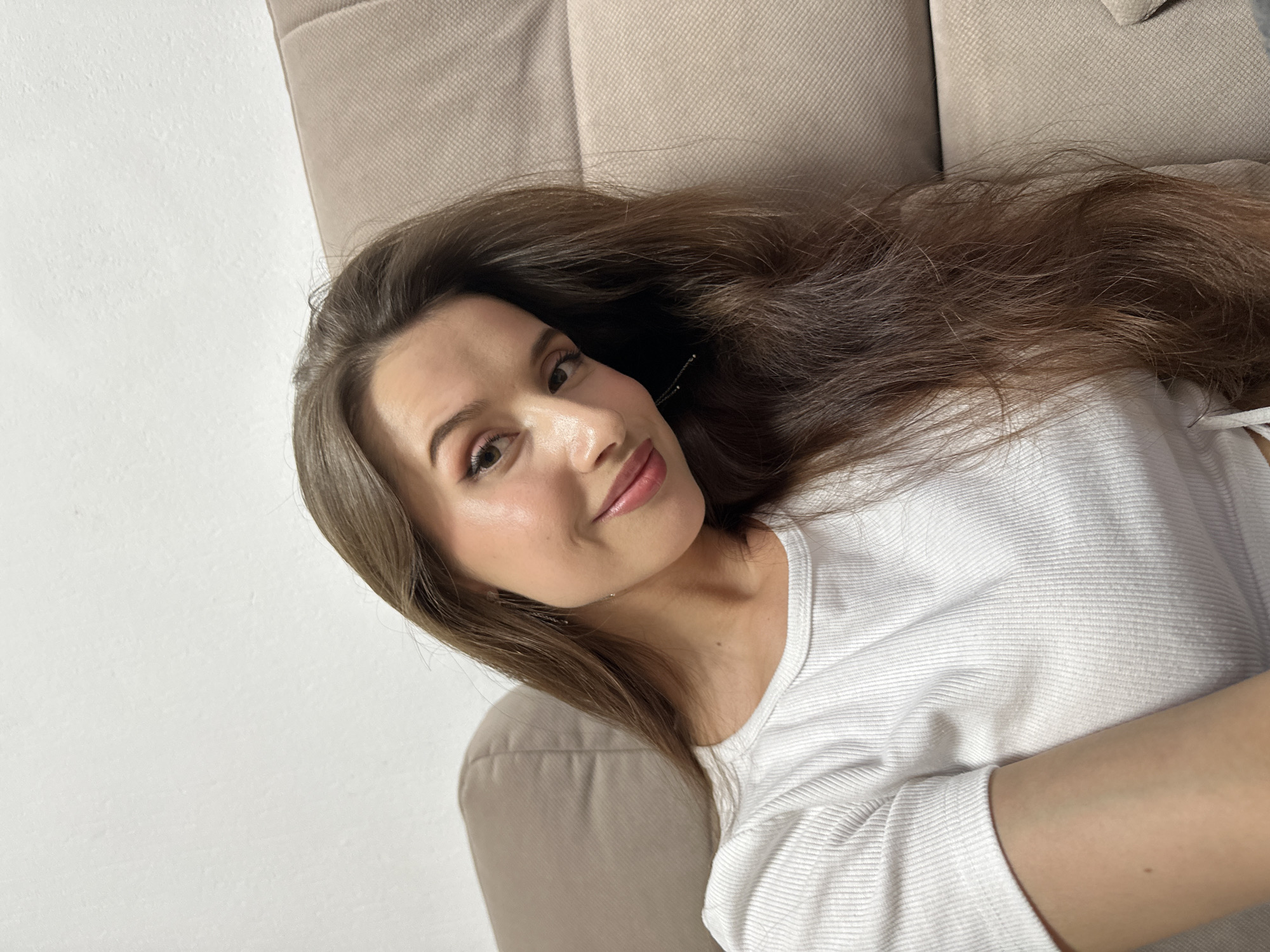 A young woman is lying on a beige sofa in what appears to be an indoor setting. She is wearing a white textured long-sleeved shirt. Her long, brown hair is spread out on the sofa. She has fair skin and is looking at the camera with a slight smile. The sofa is situated against a light-colored wall. There is no immediate information to suggest the photo was taken in Yekaterinburg, Russia.