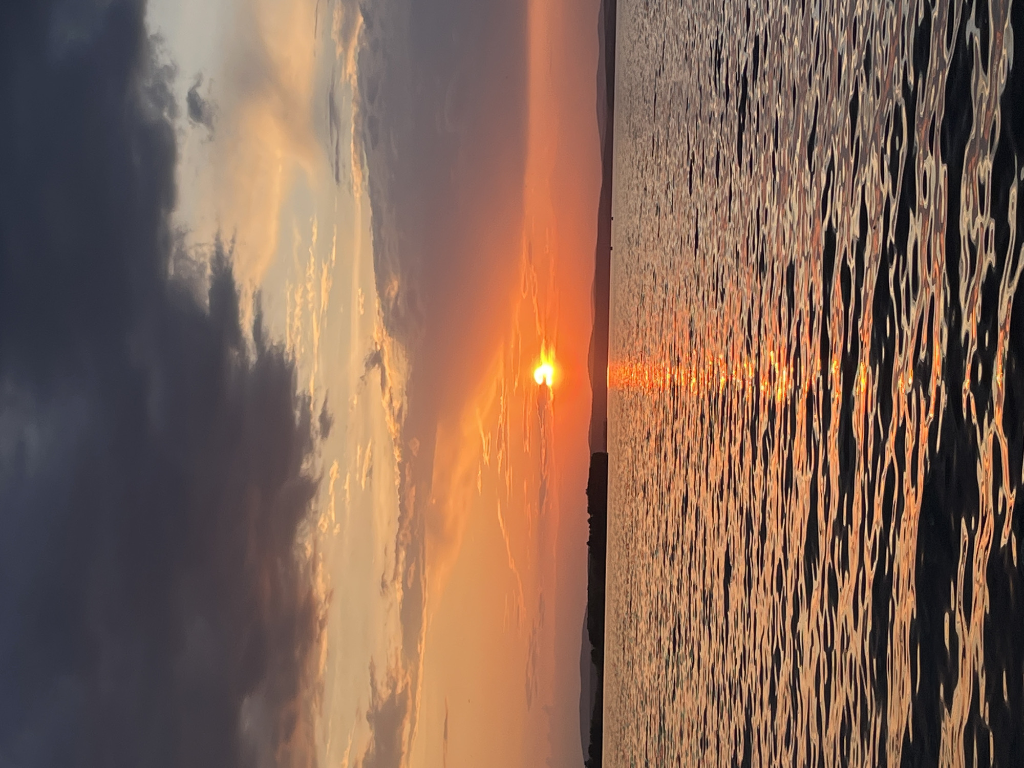 The image depicts a sunset over a body of water. The sun is positioned near the horizon, reflecting off the surface of the water. To the left, there are dark clouds and lighter, golden-toned clouds illuminated by the setting sun. A dark landmass is visible in the distance between the sky and the water. The water surface is rippled, creating a shimmering effect as it reflects the sunlight. I am unable to confirm the exact location and can only suggest that if the photo was taken in Yekaterinburg, Russia, it would be near the Iset River or Lake Shartash.