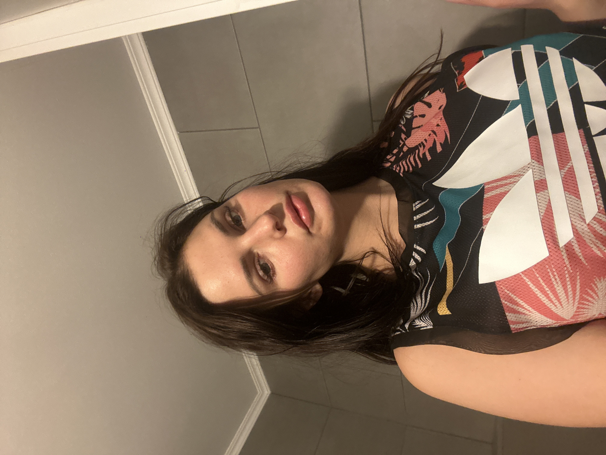 A low-angle, close-up indoor photograph shows a young woman looking down and to the right. She has long, dark brown hair and fair skin with rosy cheeks. She is wearing a black mesh tank top with a colorful abstract pattern featuring large white Adidas logos, pink palm fronds, and teal accents. The background appears to be a bathroom with gray tiled walls and white trim. The lighting is dim, casting shadows on her face.