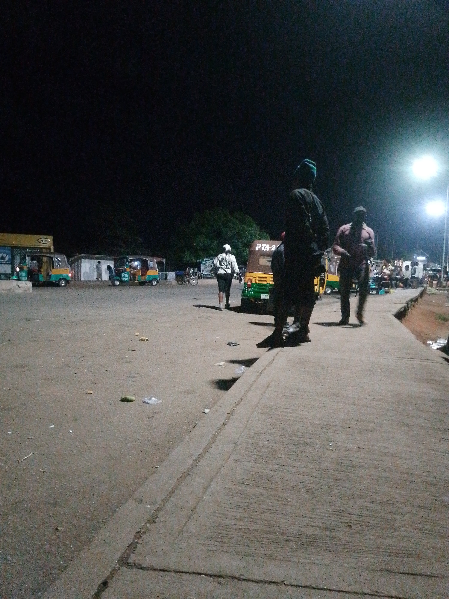 The image depicts a nighttime scene at what appears to be a transportation hub or market area. Several people are present, some standing and others seemingly engaged in activity. In the foreground, two individuals are visible from behind, one wearing a dark shirt and a green cap, and the other a lighter colored shirt. Further back, another person in a white top and dark pants is walking away from the camera.
The scene is illuminated by artificial lighting, possibly from streetlights, creating a contrast between the bright lights and the dark sky. Several three-wheeled vehicles, often used as taxis or for transport, are parked in the area. One of these vehicles has the text "PTA 2" visible on its side. Another vehicle has the word "HTD" visible on its signage. There are also some structures that look like stalls or small buildings in the background, suggesting a commercial or transport gathering point.
The ground is paved, and there is some litter scattered around, including plastic wrappers and what appear to be fallen leaves or fruit. The overall atmosphere seems busy, with people moving about. The time of day is clearly nighttime due to the dark sky and the use of artificial lighting. The weather appears to be clear, as there are no visible signs of rain or fog. The location cues are minimal, but the presence of multiple three-wheeled vehicles and market-like structures points towards a lively urban or semi-urban setting, likely in a region where these types of vehicles are common for public transport.