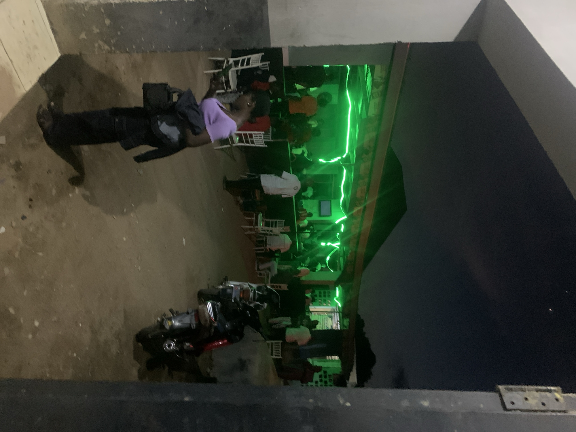This image captures a nighttime outdoor gathering in New Stadium, Nigeria. The scene is dominated by an elevated, open-air structure illuminated with bright green LED or neon-like strip lights, serving as a social area.
In the immediate foreground, a woman is positioned slightly left of center, her back partially towards the viewer. She is attired in a light purple long-sleeved top, dark trousers, and dark sandals. She carries a black handbag in her left hand and a dark jacket draped over her left arm. Her right hand appears to be holding a small, dark object.
Within the green-lit structure, approximately 10 to 15 individuals are present. Several people are seated at white plastic tables with matching white chairs, while others are standing and socializing. One individual in a white shirt and dark trousers is visible standing towards the right side of the illuminated section. Another person wearing an orange top is visible standing on the left side of the green-lit area.
The ground leading up to the elevated structure is unpaved, appearing as dirt or light-colored gravel. Two motorcycles are parked on the right side of this unpaved ground; the motorcycle closer to the viewer is predominantly dark with red accents, while the one behind it is lighter in color.
The left side of the image is framed by a light-colored building or wall structure. The right side is framed by a dark, vertical structural element that has a metallic, hinged bracket-like object attached to it in the upper portion. The night sky above is dark, showing a few faint light sources, including two distinct, small reddish-orange points in the upper right quadrant. A dark treeline or distant structure is visible against the horizon in the background.