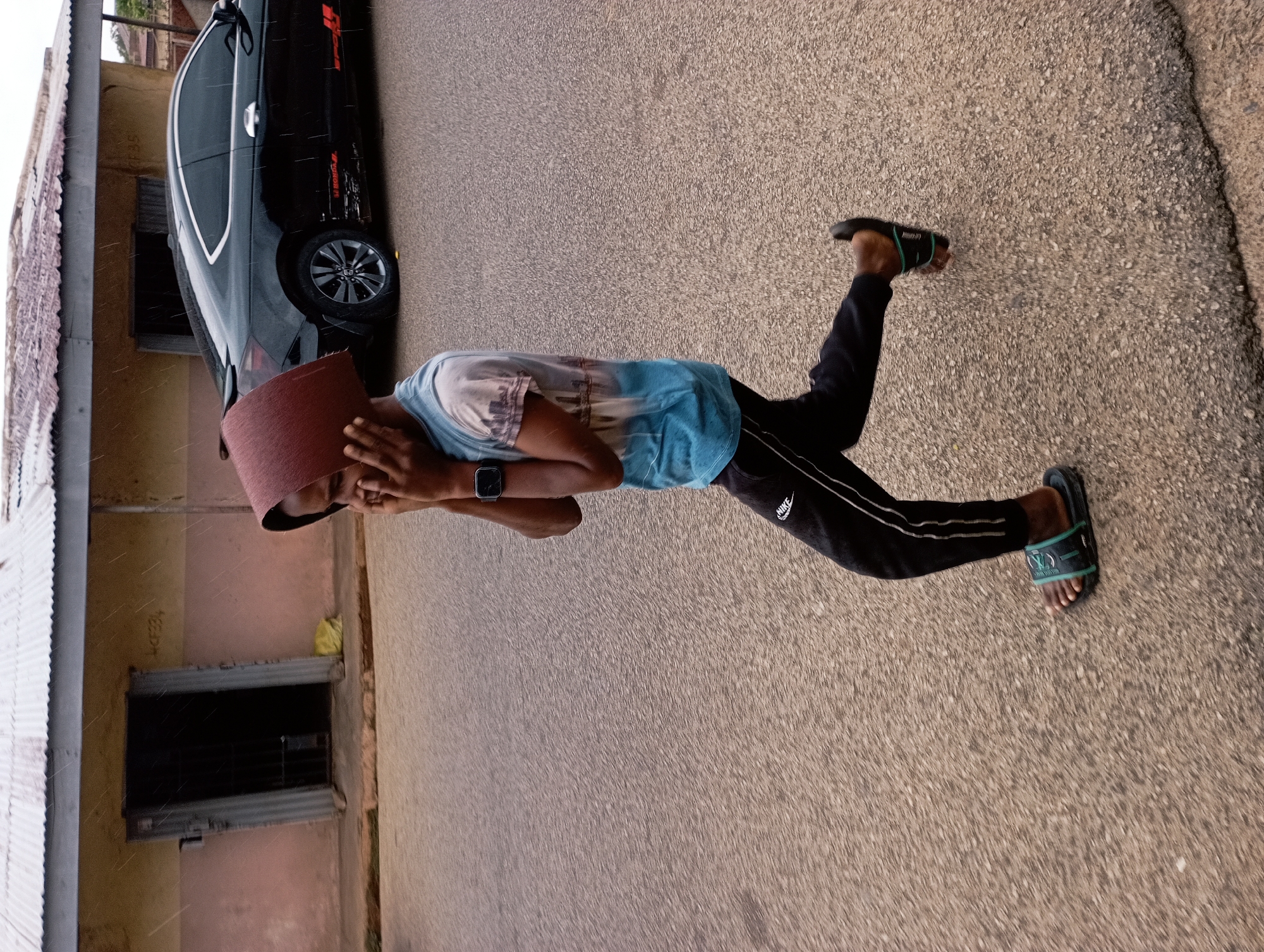 A young person, wearing a blue t-shirt with a graphic print and black track pants with white stripes, walks along a paved road. The person has a brown, curved object, possibly a hat or a basket, placed over their head and is touching their face with their hands. They are wearing dark-colored slides with green straps. In the background, a black car is parked in front of a light-colored building with a corrugated metal roof. The building has a dark-colored doorway with metal bars. The ground is asphalt, and there are visible specks of rain falling. The scene appears to be a street in Jos, Nigeria, possibly during a light rain shower.