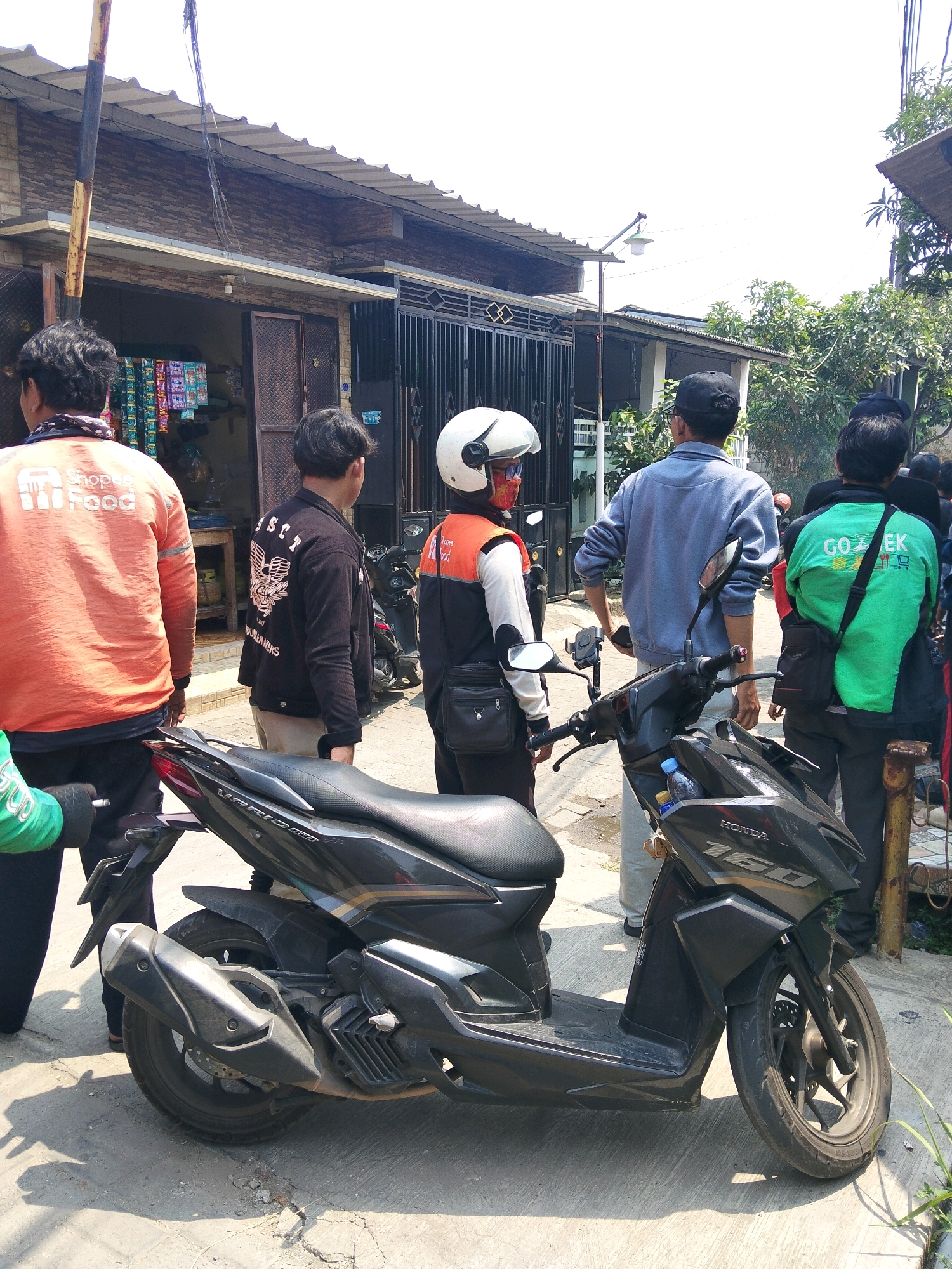 In the city of Rajegmulya, Indonesia, a group of individuals stand near a small storefront. The foreground is dominated by a black Honda Vario 160 scooter, parked on a concrete surface that shows signs of wear and cracks. Behind the scooter, a person wearing an orange Shopee Food jacket is visible, with another individual in a black jacket with a graphic design on the back standing to their right. Further to the right, a person in a light blue jacket and a person wearing a green Gojek jacket are also present. The storefront behind them appears to be a small shop with shelves stocked with various goods, and a metal gate is visible to the right of the storefront. The environment suggests an outdoor urban setting with buildings and trees in the background under a bright sky.