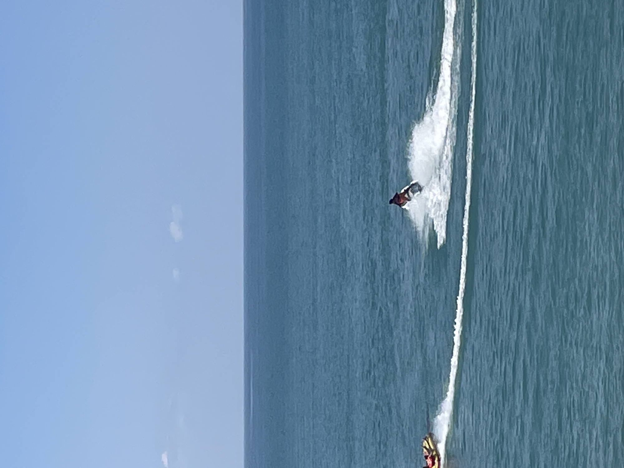 The image shows a bright, clear day with a vast expanse of blue ocean meeting a pale blue sky, separated by a thin, dark horizon line. The water is a deep teal color, with gentle ripples across its surface. In the mid-ground, a single person is riding a jet ski, leaving a white spray of water in their wake as they move from left to right. Further to the left, also in the mid-ground, a yellow inflatable tube with two people is being towed, creating a similar white trail on the water. The scene appears to be set at sea, likely in a recreational area. There is no visible indication of the city of Giachrypsh, Abkhazia in this image.
