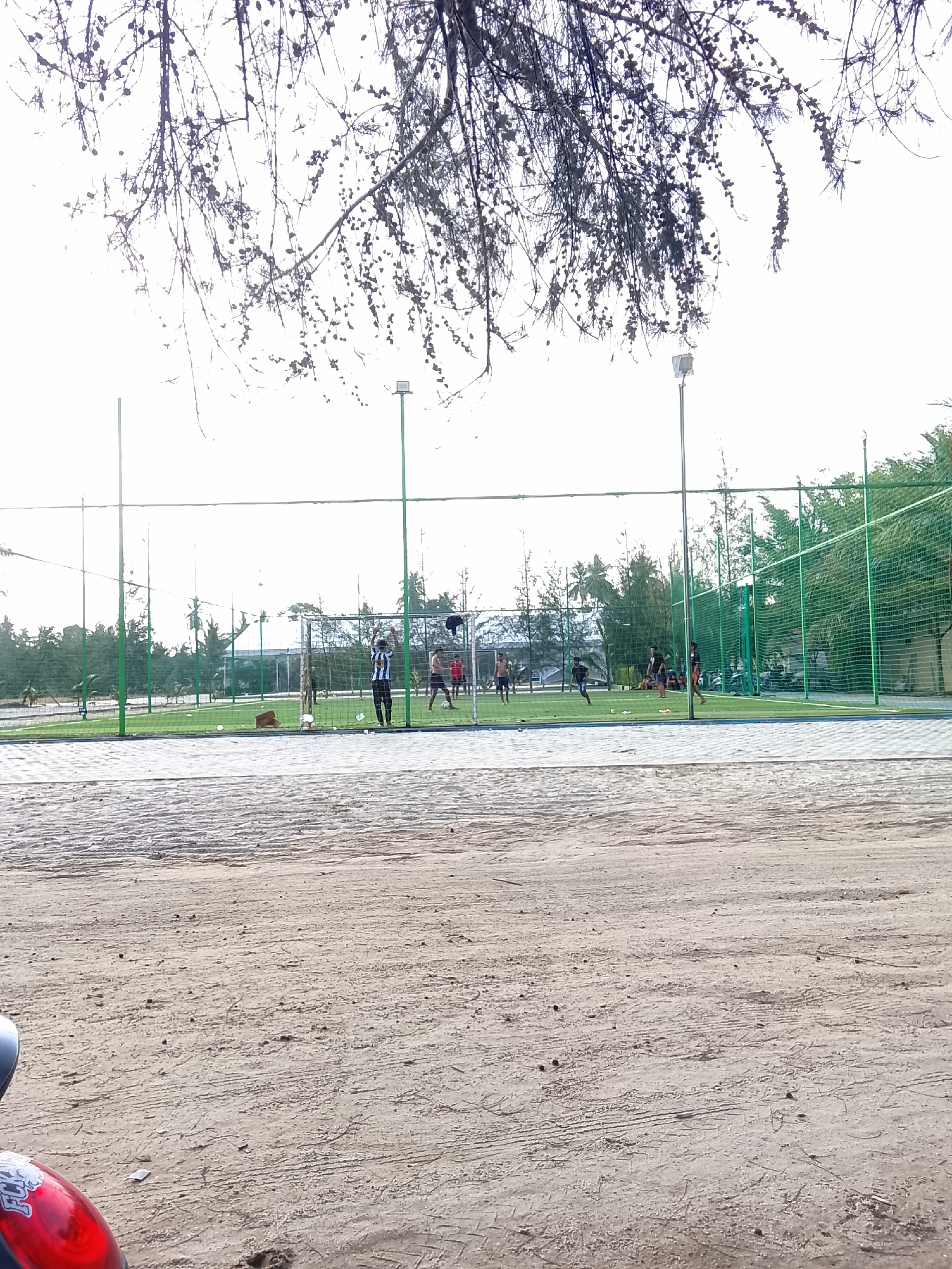 The image captures a scene on a sandy area in front of a fenced-in, artificial turf sports field. The field appears to be a soccer or futsal court, with goalposts visible. Several individuals are present on the field, actively engaged in a game, with some running and others near the goal. A person in a striped shirt stands near the goalpost. The field is surrounded by a green net enclosure, and tall green poles with lights are positioned around it. Beyond the field, a line of trees, including palm trees, forms a backdrop, with some buildings and what appear to be parked vehicles or motorcycles visible in the distance. The foreground is dominated by a sandy, textured ground with some debris. The overall lighting suggests an outdoor daytime setting, possibly overcast due to the bright sky.
