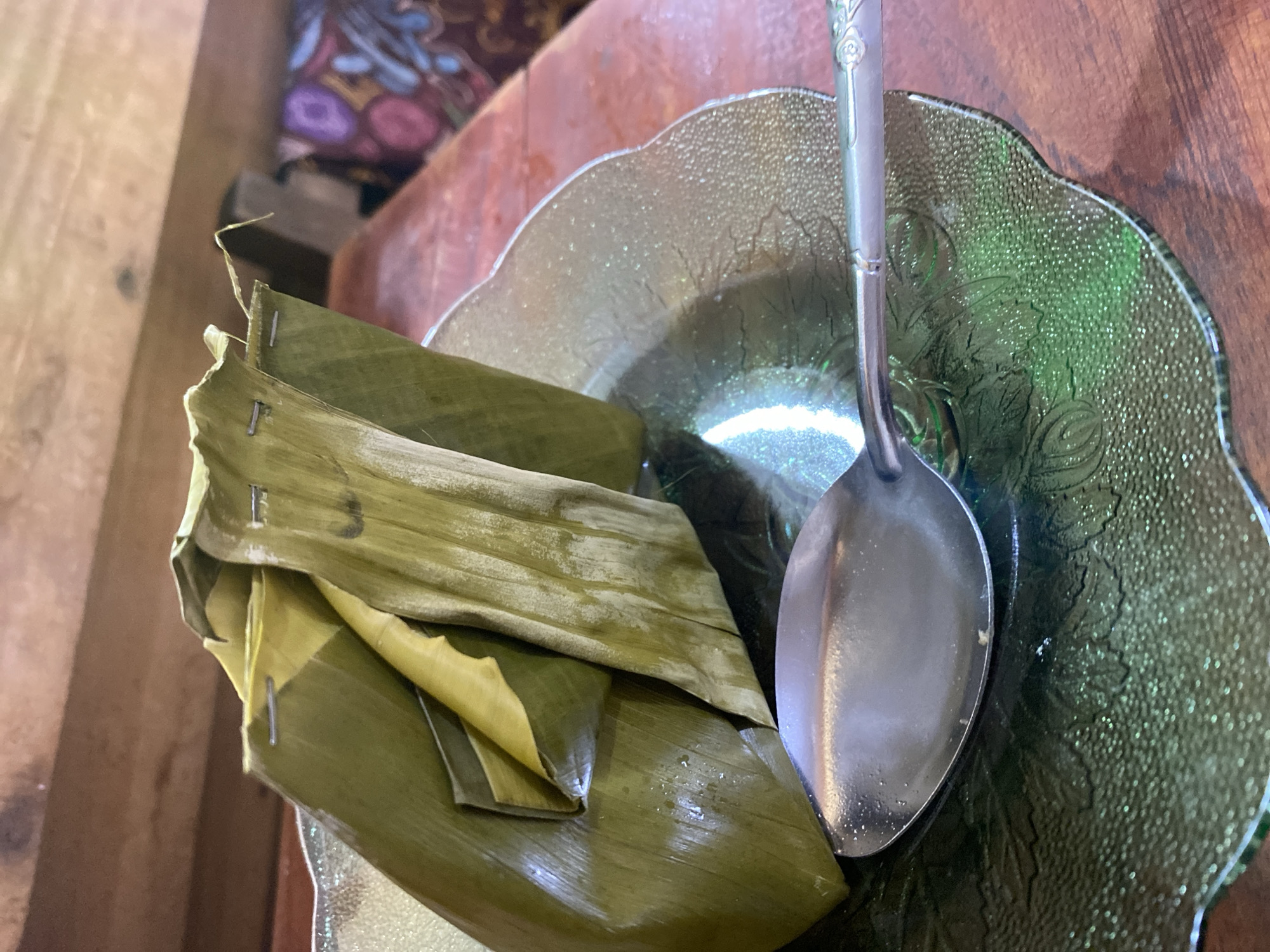 A close-up, low-angle shot shows a dish wrapped in a banana leaf, placed on a green glass plate. The banana leaf is secured with small staples. A spoon rests in front of the dish, on the plate. The plate has a raised, textured floral pattern and is set on a wooden surface. The background is blurred, but appears to be an indoor setting with wooden beams and a patterned fabric. No people are visible in the image. The scene depicts food preparation or serving, likely in a home or traditional eatery in Ta', Indonesia.