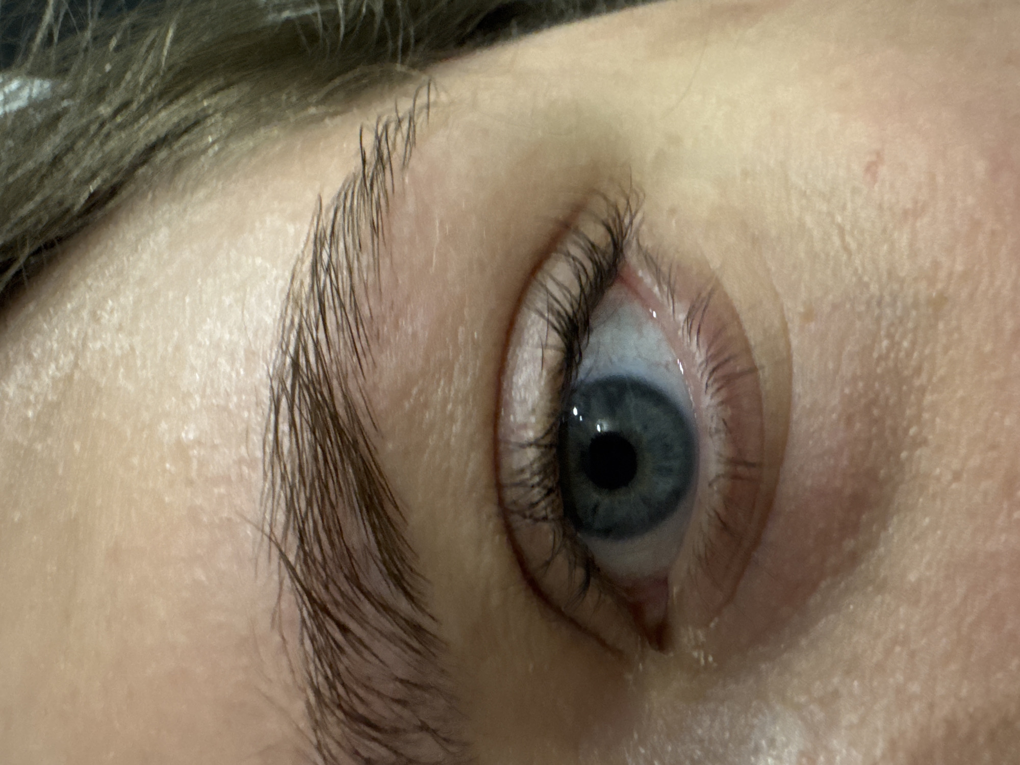 The image is a close-up, eye-level shot of a human eye with a blue iris and dark lashes. The skin surrounding the eye appears fair with some visible pores and slight redness. There are dark, bushy eyebrows visible above the eye and strands of brown hair to the right. The background is dark and out of focus, obscuring any environmental details. There is no information or visual evidence within the image to suggest the location in Saratov, Russia.
