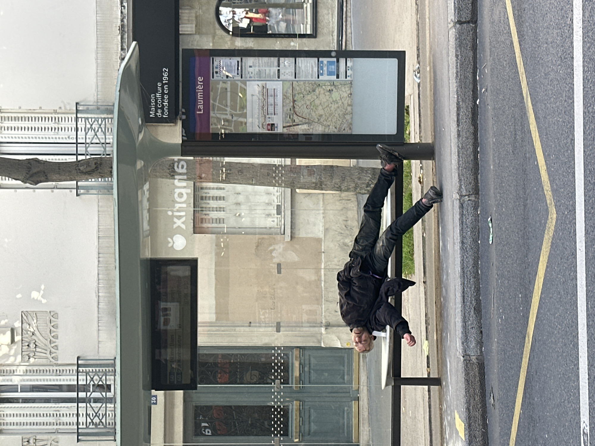 A man is lying on a bus stop bench on a city street. The street is divided by a yellow line next to a white line. The bus stop has signage with the word "Laumière" and a map. On the glass of the bus stop is an advertisement that says "Xingris Maison de coiffure fondée en 1962." The number "30" is visible on a plaque attached to the building near the bus stop, as well as "60 B'd Poissonniere. 60 R. de Montmartre". The street is lined with buildings. The image is rotated 90 degrees clockwise.