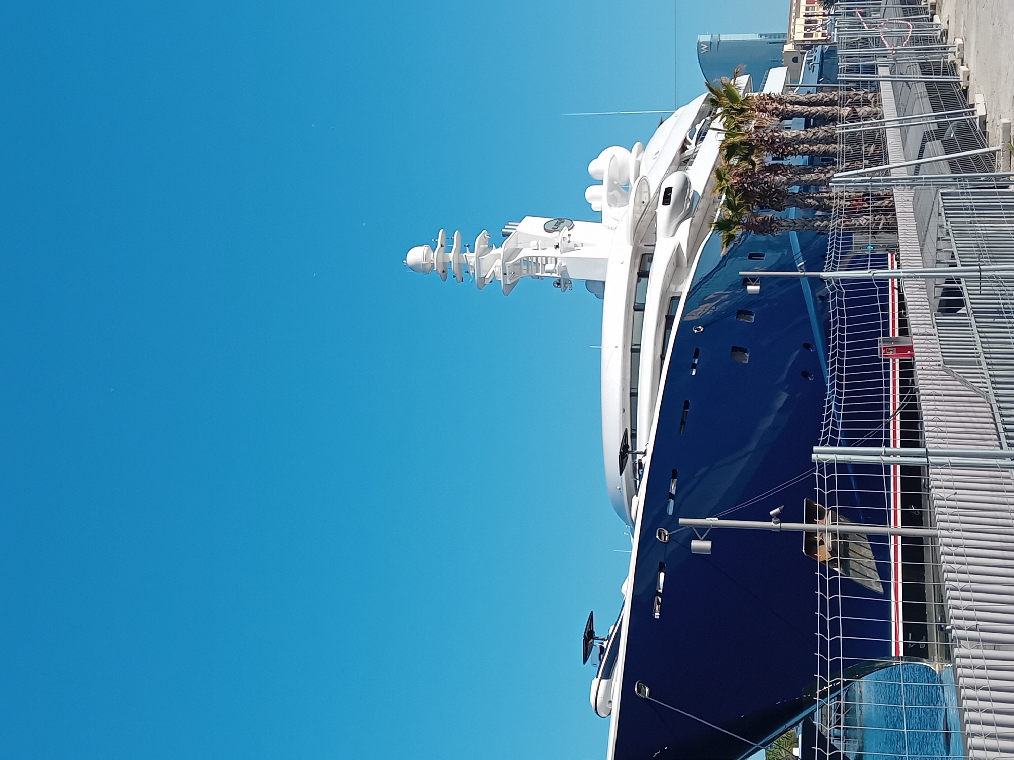 A large, modern superyacht is prominently docked in a marina under a clear, bright blue sky on a sunny day in Barcelona, Spain. The yacht occupies the right side of the frame, showcasing its sleek design with a deep, dark blue hull and a multi-tiered white superstructure. Various navigation and communication equipment, including radar domes and antennas, are visible on its mast and upper decks.
In the mid-ground, a row of tall palm trees with mature, textured trunks and green fronds stands between the yacht and a grey metal fence. The fence, possibly a temporary barrier for the dock, runs across the foreground, creating a visual separation between the viewer and the superyacht. Part of the yacht's bow, including a stylized anchor, is visible behind a section of the fence.
In the far background, beyond the palm trees and yacht, faint architectural details of distant buildings are visible, including a prominent "W" logo on one structure, likely indicating a W Hotel. The overall scene suggests a vibrant, luxurious port environment bathed in clear daylight, with no specific activity beyond the vessel being moored.