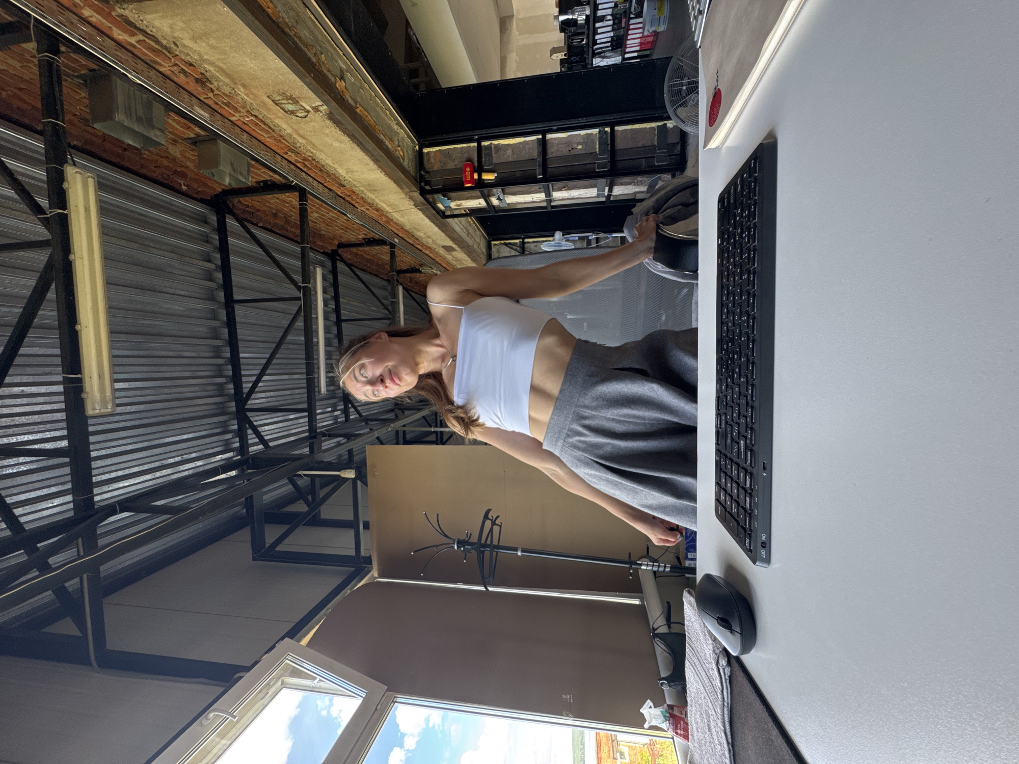 A young woman is captured in a full-body shot, posing dynamically in what appears to be a spacious, industrial-style room. She is wearing a white crop top and grey wide-leg pants. Her arms are outstretched, with one hand resting on a large black object, possibly a large hat or helmet. She is looking directly at the camera with a neutral expression.
The background reveals a high ceiling with exposed brickwork and metal beams, suggesting a renovated industrial space or loft. Large metal roll-up doors are visible, hinting at a workshop or storage area. Natural light streams in from what seems to be a large window or opening, casting a bright ambiance. A modern desk with a keyboard and mouse is positioned to the right, indicating a workspace within the larger room. A coat rack stands in the mid-ground.
The overall scene suggests a casual moment of personal expression within a unique, perhaps creative or work-related, environment. The lighting implies it is daytime, possibly late morning or afternoon, given the brightness. The image seems to be taken indoors, with hints of an outdoor view through the window.