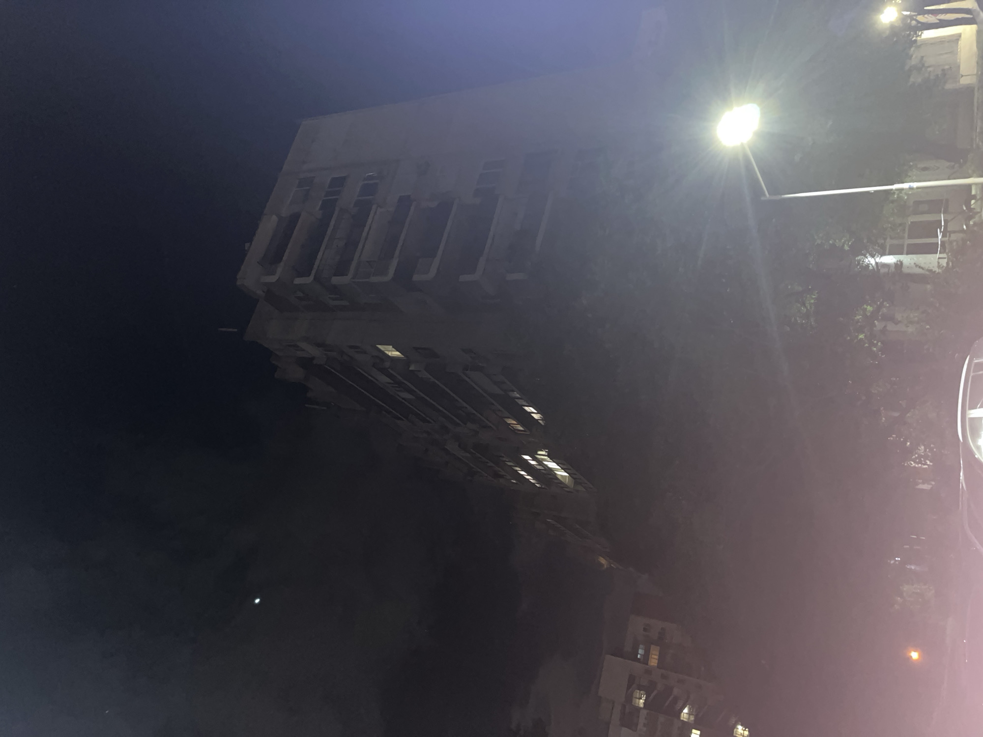 Here is a description of the image:
The image captures a nighttime scene, likely in an urban environment. A tall, multi-story building dominates the left side of the frame. It features a series of vertically-oriented recessed structures that could be balconies or window bays. The building appears to be illuminated by lights both inside and from exterior sources. To the right, a streetlight is prominently displayed, with a halo effect surrounding the bulb. Additional buildings are visible in the distance, with lit windows suggesting residential or commercial activity. The presence of trees along the street further enhances the urban setting. A vehicle is visible on the right side of the scene. The overall impression is one of a city street at night, with a mix of architectural elements, artificial lighting, and natural elements.
