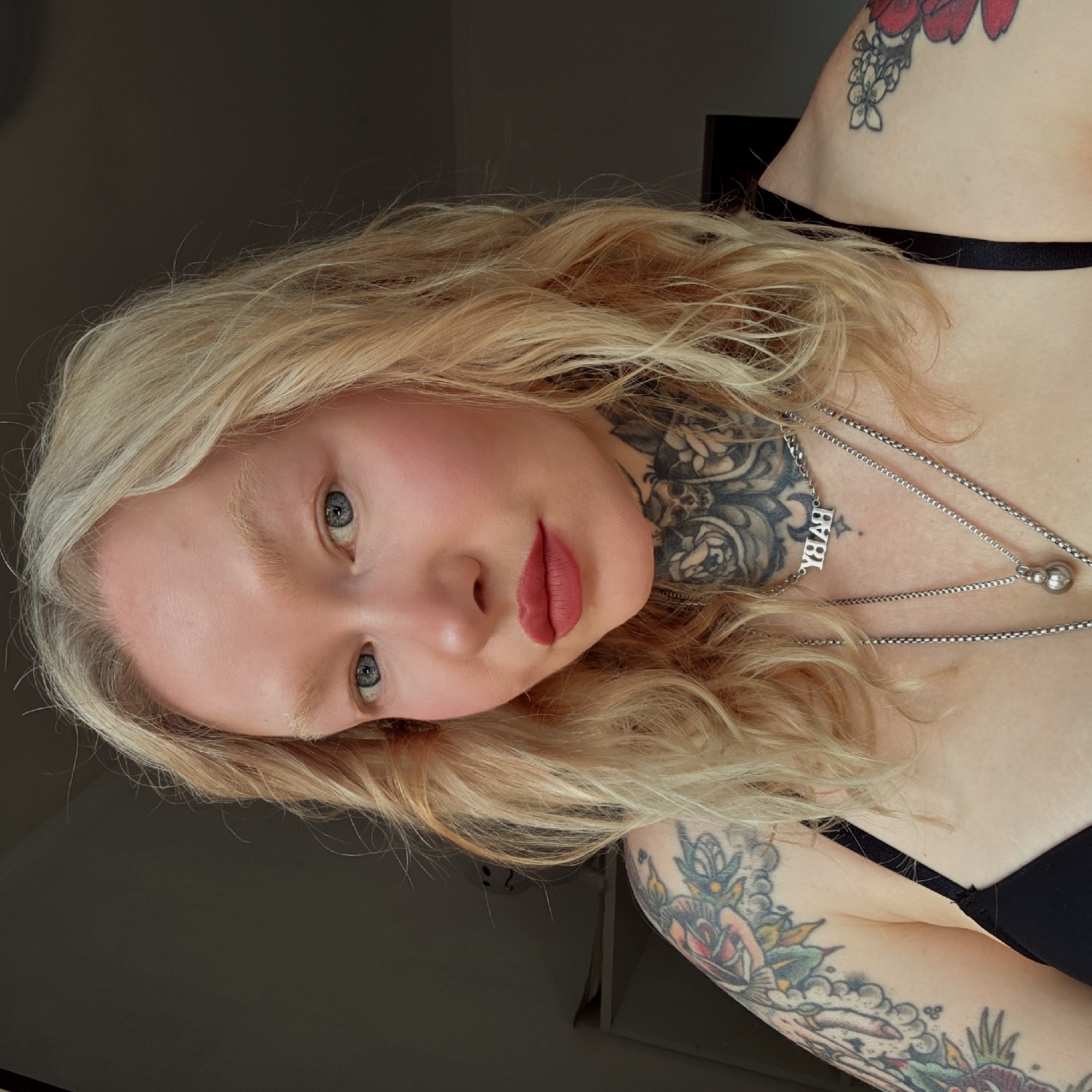 A fair-skinned person with blonde hair, blue eyes, and red lips is pictured from a low-angle perspective. The person has numerous tattoos, including floral designs on their upper arms and a detailed tattoo on their neck that appears to include skulls and roses. They are wearing layered silver necklaces, one with the word "BABY" in block letters. They are wearing a black top with thin straps. The background is dark and indistinct, not providing any indication of the location in Saint Petersburg, Russia.