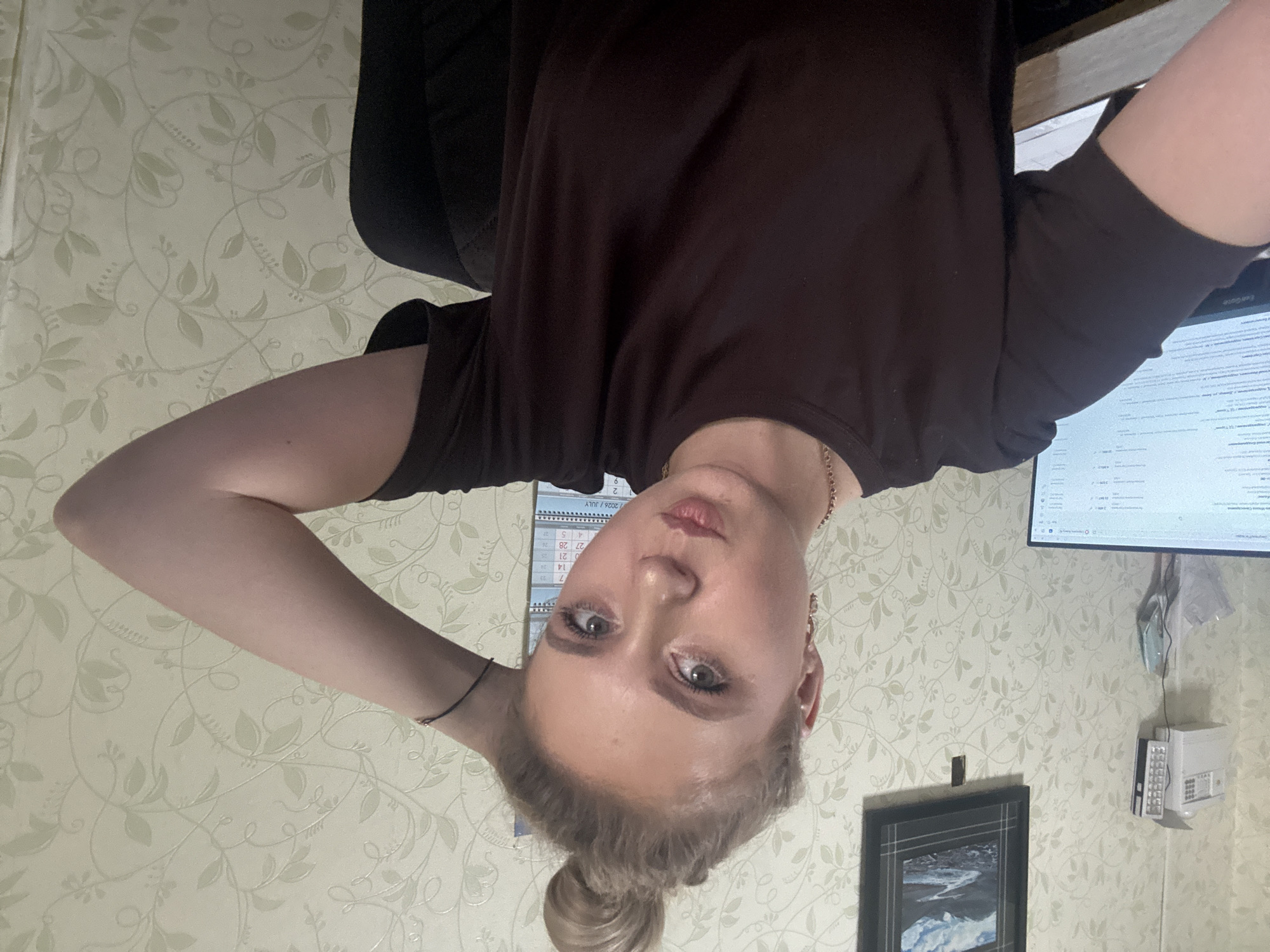 A young woman with blonde hair tied in a bun is looking at the camera with a playful expression, resting her right arm on her head. She is wearing a dark brown t-shirt and a gold necklace. Behind her, a computer monitor displays a spreadsheet, and a wall with floral wallpaper is visible. A framed picture of a mountain landscape hangs on the wall, along with a white emergency light fixture and a calendar. The scene appears to be an indoor office or home setting.