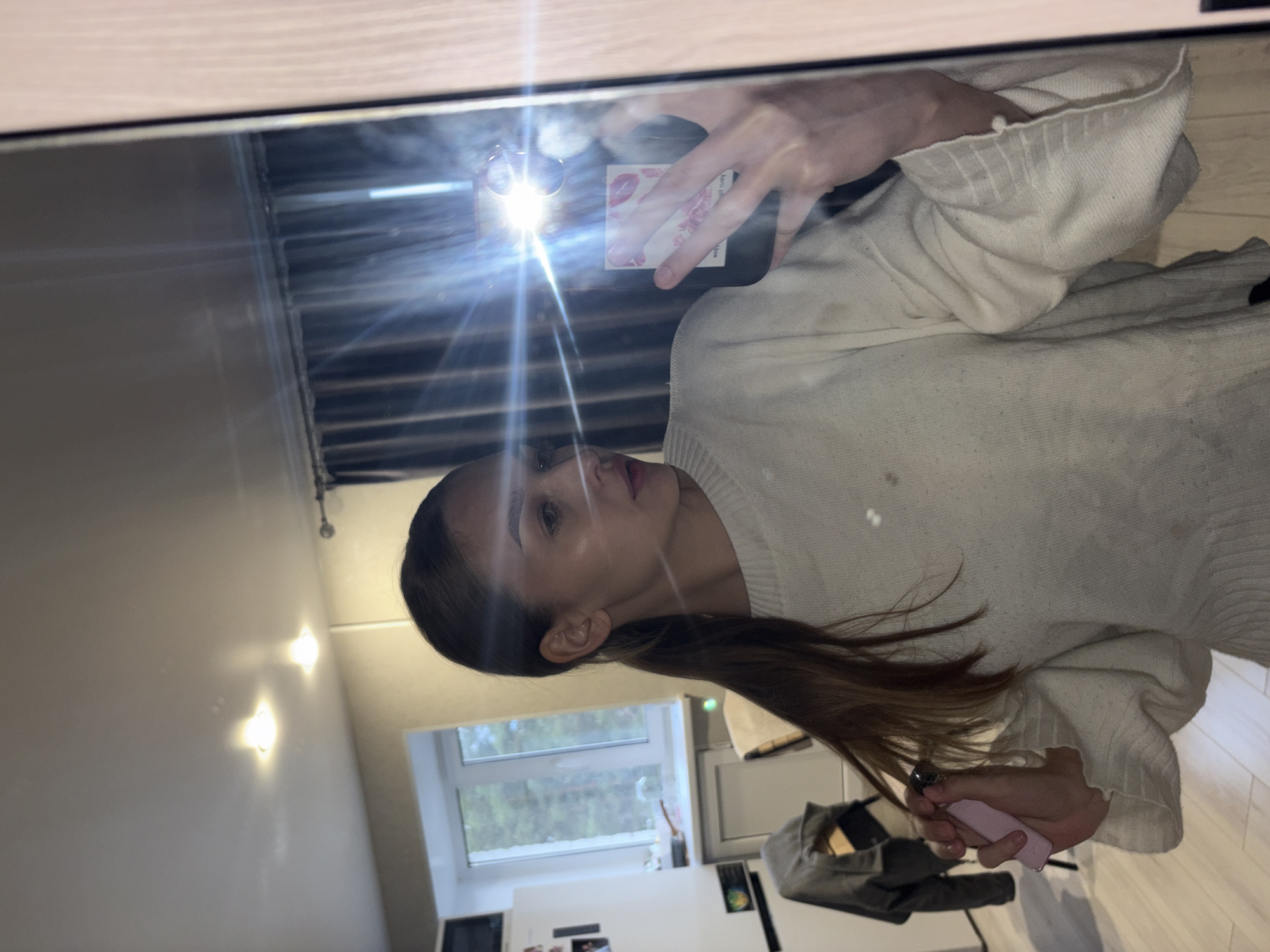 A person in a light-colored sweater is taking a mirror selfie in what appears to be an apartment or home, likely in Chaikovsky, Russia. A portion of the interior of the apartment is visible in the background, including a window overlooking some trees, kitchen appliances, and overhead lighting. The person is holding a phone up to the mirror. They are also holding a lighter in their hand, which is being reflected in the mirror.