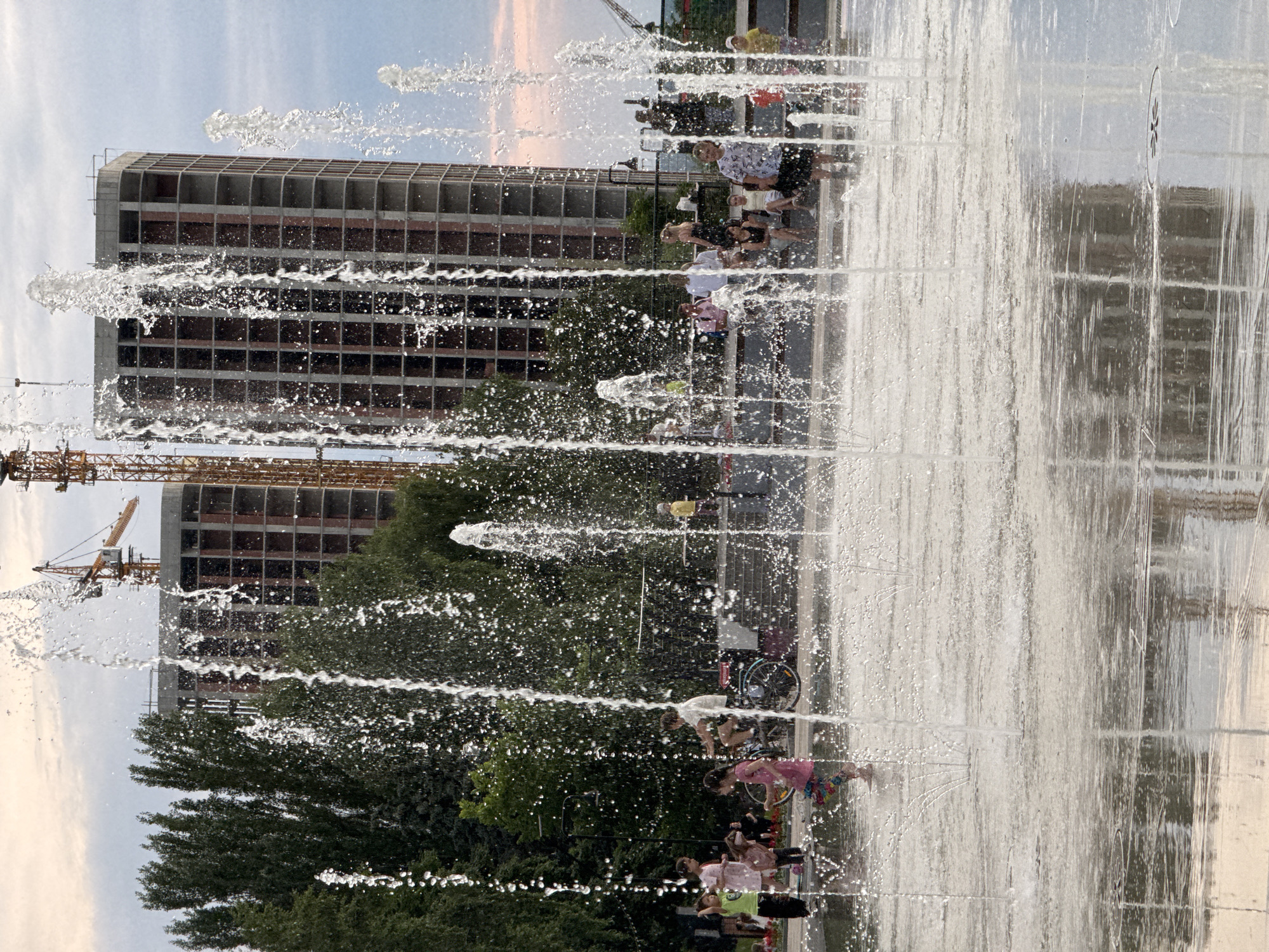 The image depicts a contemporary urban park scene with a water fountain play area in the foreground. Several children are actively playing in the shallow water, with some running and others standing. In the middle ground, a group of people are seated on a bench and a ledge, observing or resting. Behind them, a bicycle is parked. The background reveals construction sites with unfinished buildings and cranes, suggesting ongoing urban development. Tall green trees frame the scene, and the sky above is overcast with a hint of sunset colors. The location is identified as the Embankment of the Friendship of Peoples in Izhevsk, Russia.