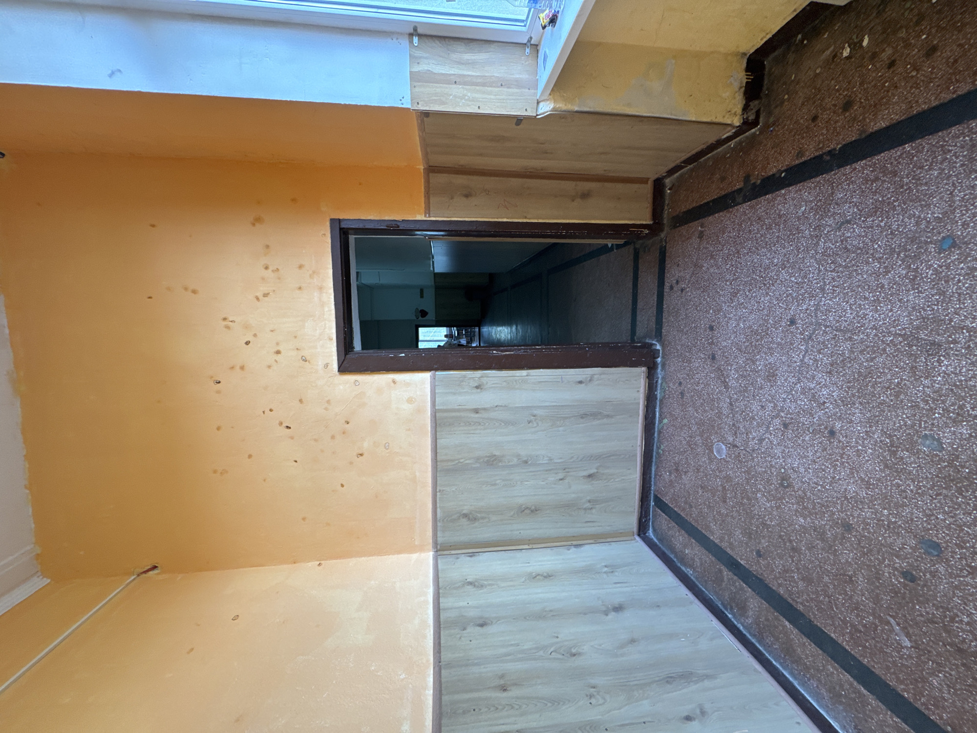 The image shows an interior space, possibly a hallway, in Buhovo, Bulgaria. The walls are painted in a dull orange, with what appears to be staining or discoloration. The floor is covered with light wood-patterned laminate flooring in one area and brown, speckled linoleum flooring in another. A doorway with a dark brown frame is visible, leading to a dark, long hallway. The room is not well-maintained, and there are signs of wear and tear. There are no people visible in the photograph.