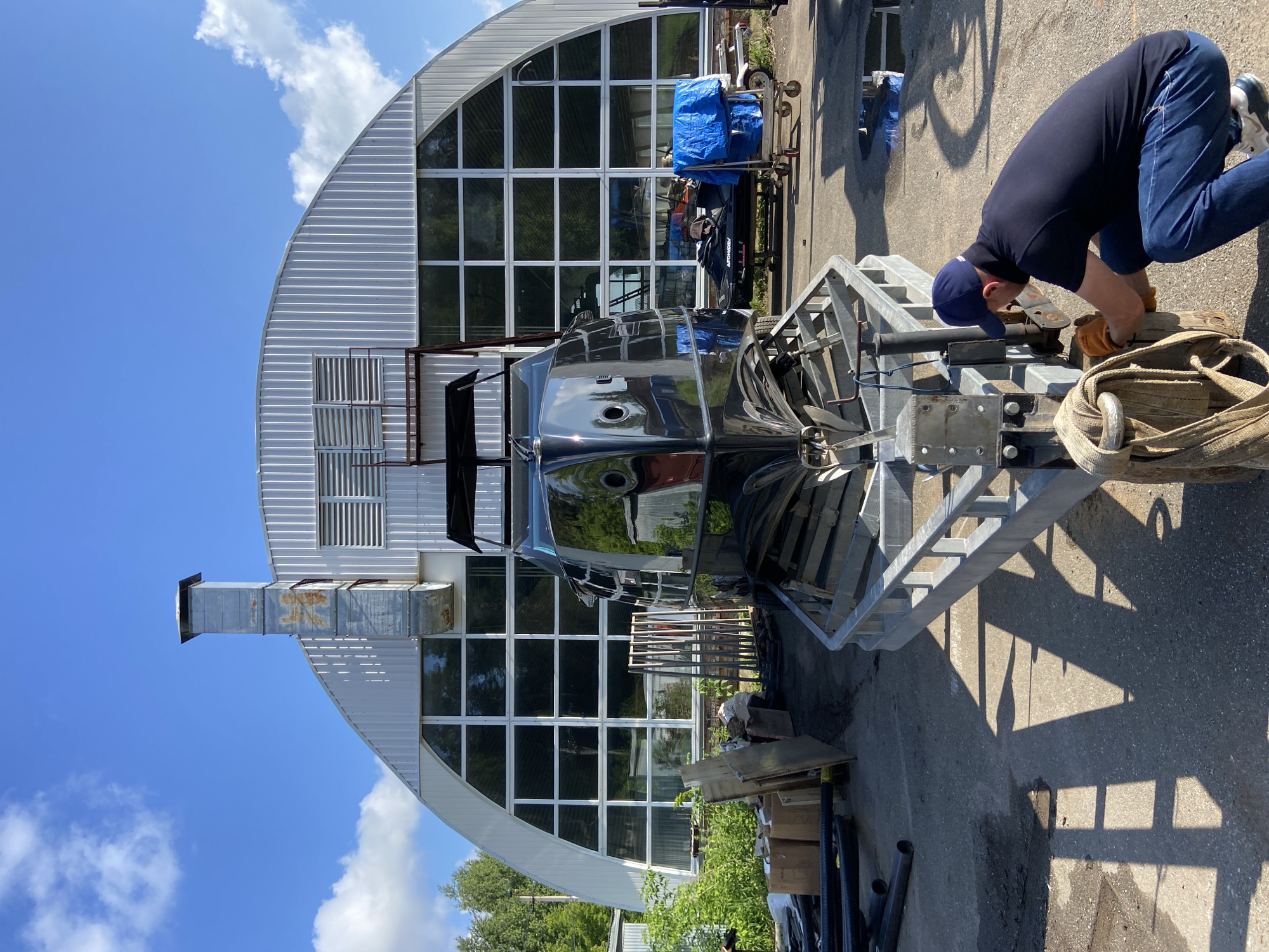 The image depicts an outdoor scene in front of a large, arched, corrugated metal building with numerous windows. In the foreground, a person wearing a dark blue cap, a dark blue polo shirt, and blue jeans is squatting and appears to be working on the mechanism of a boat trailer. The person is wearing orange gloves. The boat trailer is carrying a glossy black boat, the bow of which is prominent in the center of the frame. Various objects, including pipes, cardboard boxes, and wooden planks, are scattered around the foreground and mid-ground. A jet ski covered with a blue tarp is visible in the mid-ground, next to a wheeled cart. The ground is asphalt, with some puddles and debris. Trees and vegetation are visible in the background, to the left of the building. The sky is blue with some white clouds. The overall impression is that of an industrial or boat repair yard. No specific landmarks or indicators of the city of Votkinsk, Russia, are visible within the image itself.