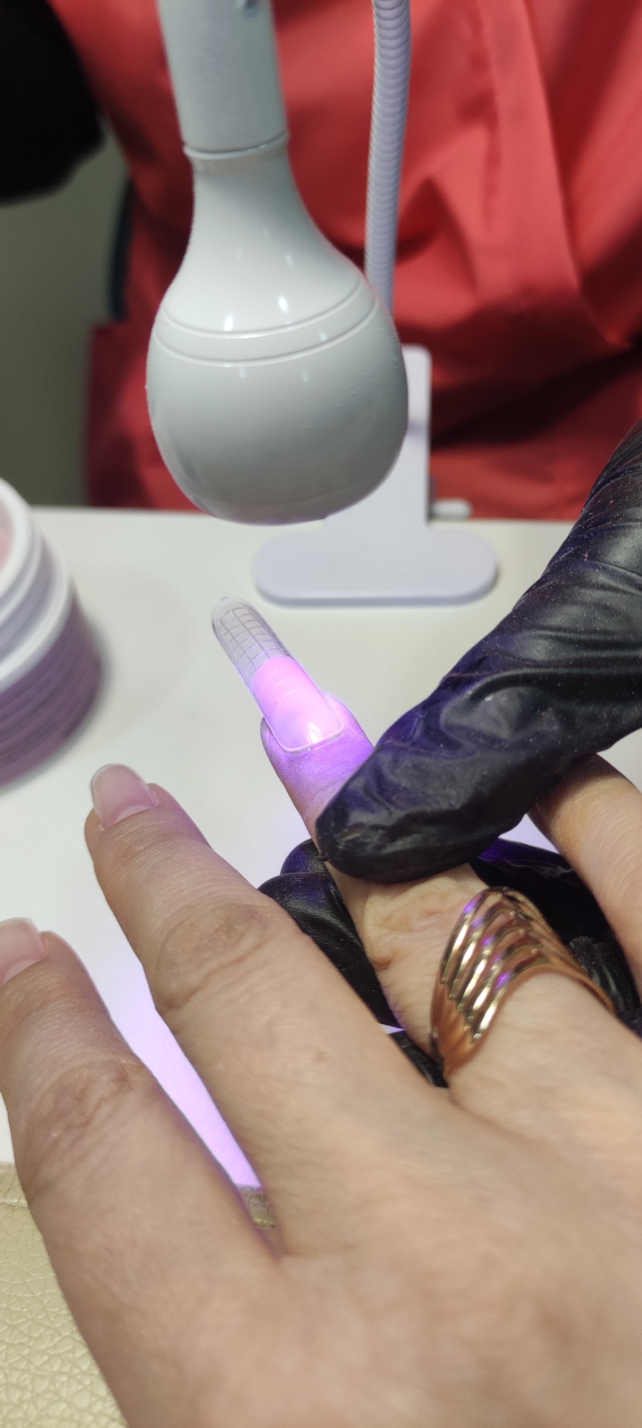 A close-up shows a person's hand with a transparent form attached to their fingernail, being held in place by a gloved hand. Above the hand, there is a white lamp. The scene appears to be indoors, with a white table surface visible. A container with stacked items is partially visible in the background. The person wears a gold ring on their finger. The location, Chaikovsky, Russia, is not deducible from the image.