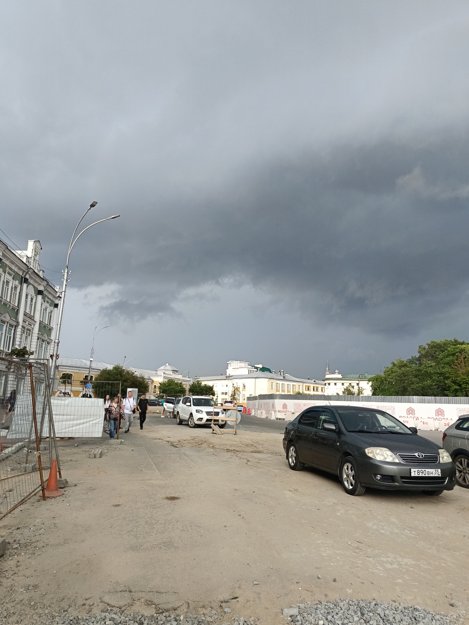 A wide-angle, eye-level, outdoor shot captures a street scene under a dark, stormy sky in an urban environment. The foreground is a dirt and gravel construction area with a bright orange traffic cone and a metal construction fence. A dark gray sedan is parked on the right side of the frame, its license plate reading "T890BH 35". In the middle ground, a group of people are walking on a paved sidewalk, with a white SUV parked behind them. The left side of the image features a green and white building with many windows, typical of historic European architecture. Further in the background, other buildings and trees are visible. The overall atmosphere is somber due to the heavy clouds.