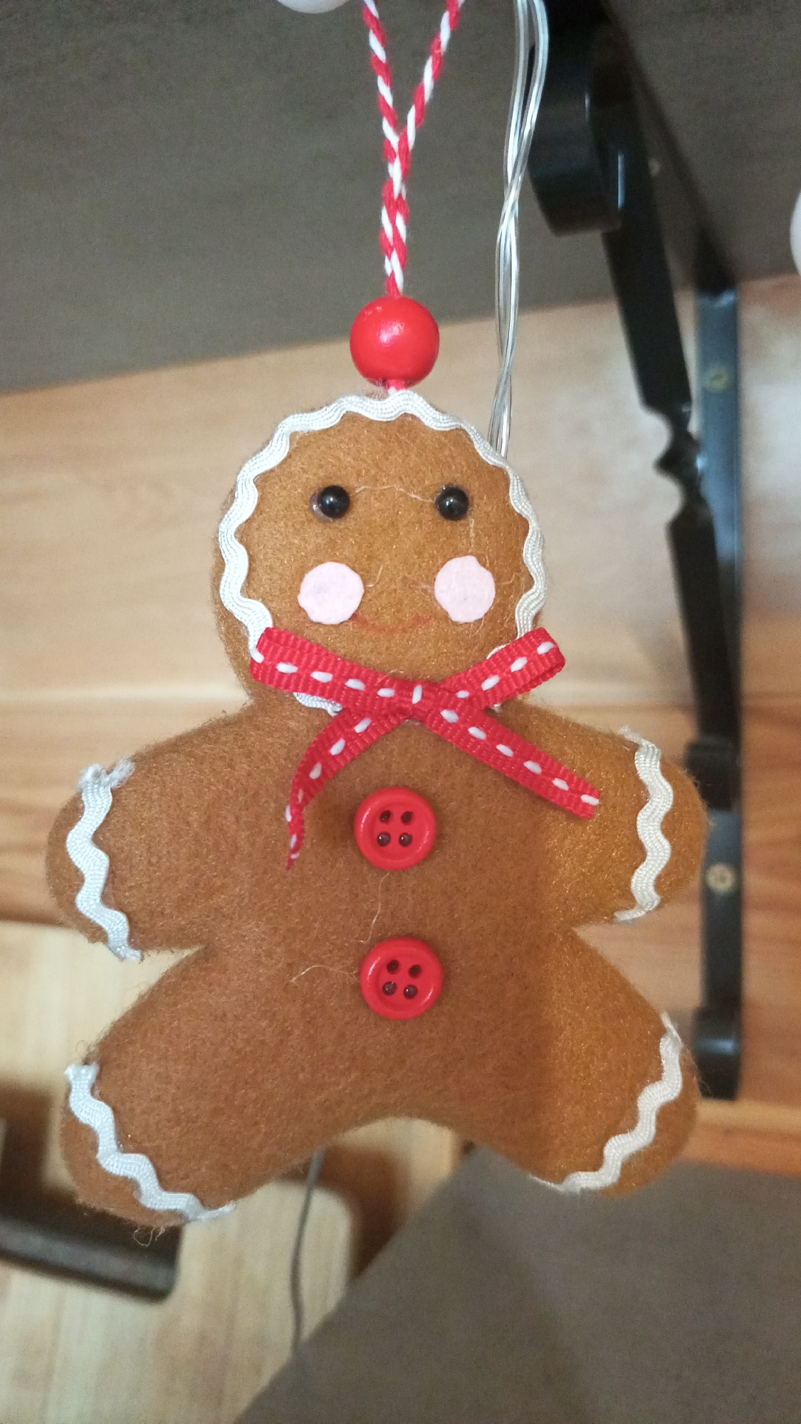 The image shows a felt gingerbread man ornament with white rickrack trim around the edges, black button eyes, pink circle cheeks, a red polka dot ribbon tied in a bow around the neck, and two red buttons down the center of the body. A red bead is attached at the top of the head with a red and white twisted cord for hanging. In the background, there is a light wood paneling and a black metal bracket with curved edges. The location of the image is not identifiable from the provided information and the image does not contain enough information to determine if it was taken in Юдино, Russia.