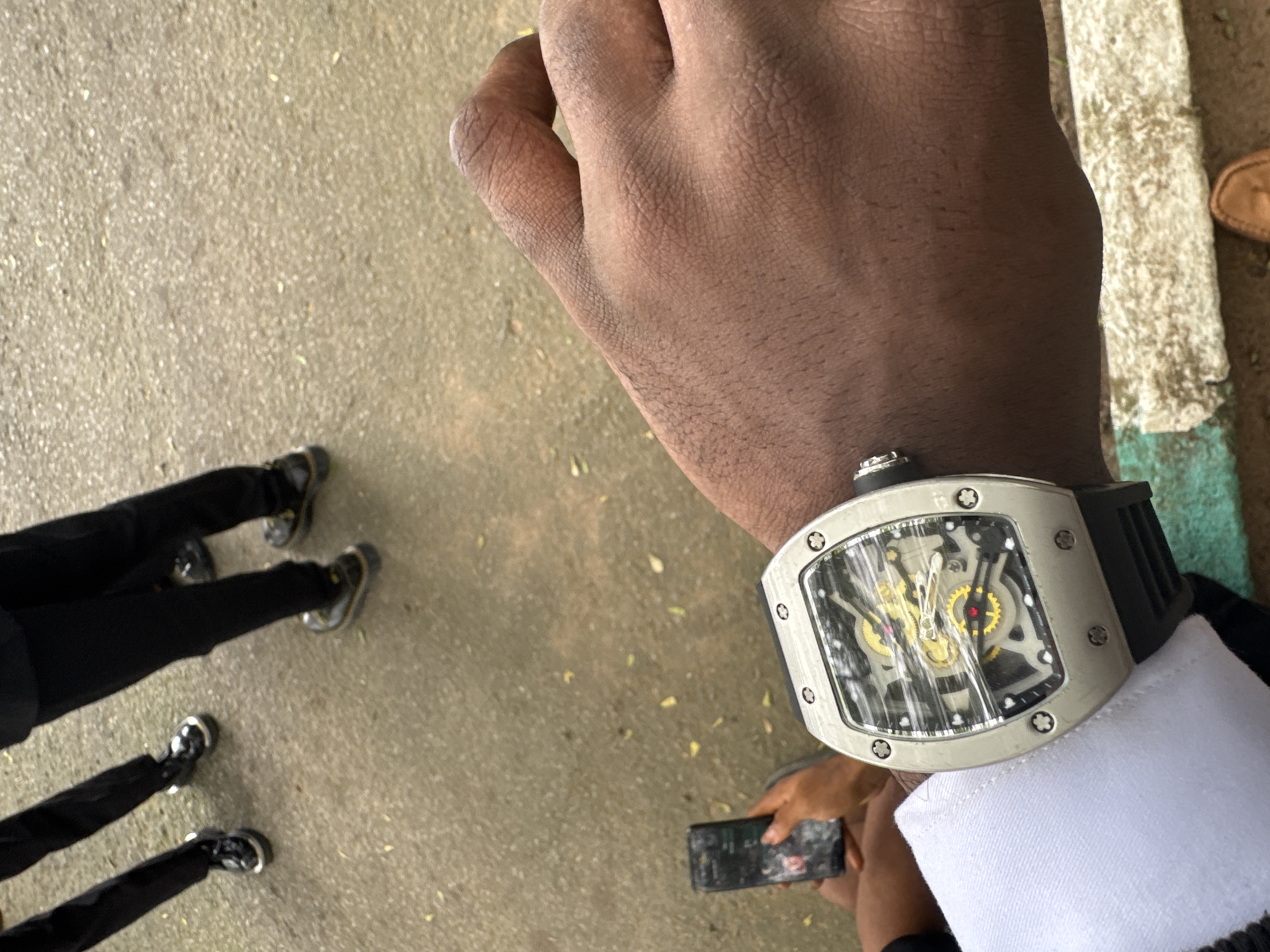 The image shows a man's wrist with a silver-cased watch. The watch has a skeletonized face with visible gears. He is wearing a white shirt with a black jacket. He is holding a smartphone. There are shoes and pants legs of multiple people visible on the left side of the image. The setting appears to be outdoors. The location is Eziobo, Nigeria.