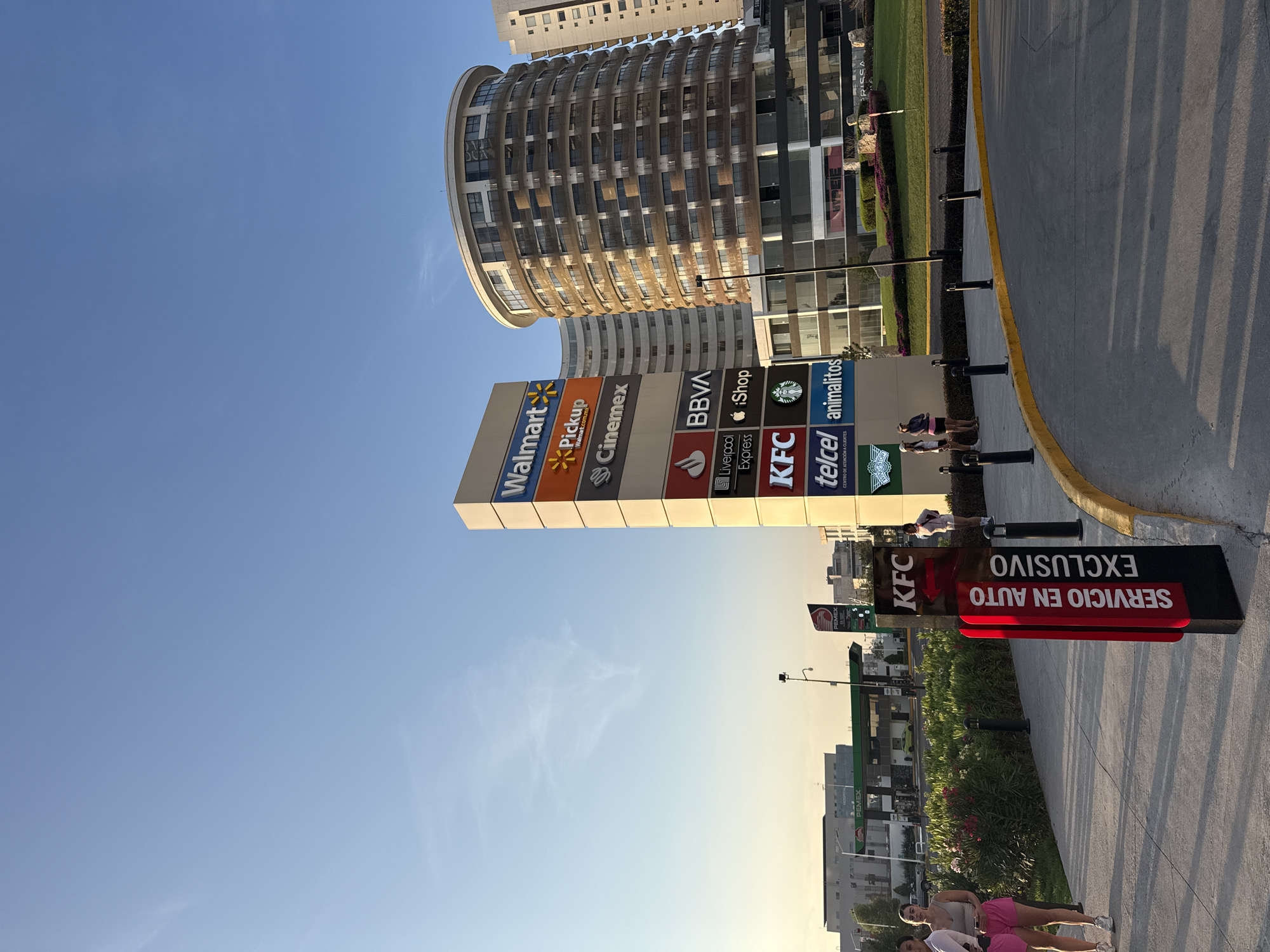 The image depicts a view of a commercial area in San Bernardino Tlaxcalancingo, Mexico. The foreground includes a prominent signpost for KFC with "SERVICIO EN AUTO EXCLUSIVO" ("Drive-Thru Exclusive") in Spanish. A group of people are visible nearby. In the middle ground, a large sign lists various businesses including Walmart, Walmart Pickup, Cinemex, BBVA, Liverpool Express, iShop, KFC, Telcel, and animalitos. Behind the signs, a modern, cylindrical building is partially visible, suggesting commercial or residential use. The background includes other buildings and infrastructure. The sky is clear and blue, suggesting daylight hours. A Pemex gas station is also visible in the background.