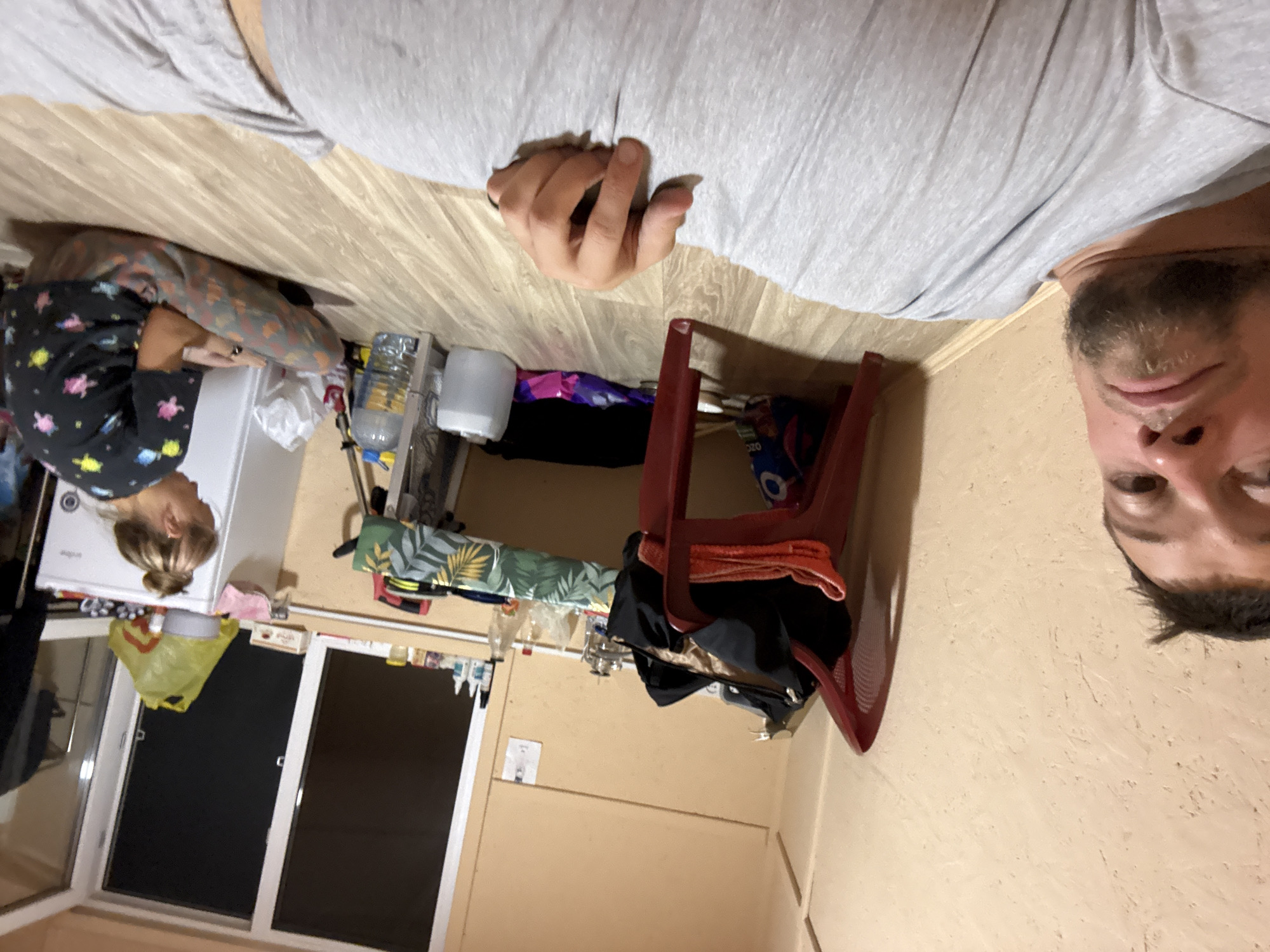 The image depicts an indoor scene, likely a modest living space in Kleshchevka, Russia. The overall ambiance is somewhat cluttered and informal. The primary subject in the foreground is a man lying on the floor, facing the camera, with his upper body and face visible. He is wearing a grey t-shirt. His expression is neutral.
In the background, a woman is seated on the floor, facing away from the camera and towards a white refrigerator. She is wearing a black t-shirt with colorful patterns and patterned trousers. She appears to be occupied with something in her hands.
The environment includes a wooden floor, beige walls, and a window with dark panes. Various objects are scattered around the room, suggesting a lived-in space. These include a red chair with a black bag and some fabric draped over it, a table with a patterned tablecloth, bottles of liquid, plastic containers, and bags. The lighting is dim, with light coming from the window and possibly an unseen light source. The scene appears to be a snapshot of everyday life, possibly in a dormitory or a small apartment.