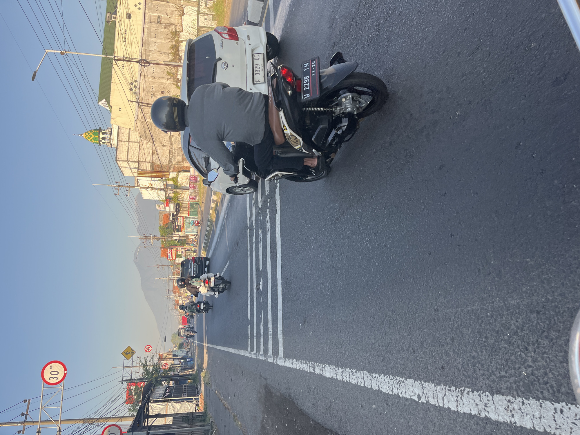 The image shows a street scene in Siring, Indonesia, during the day. The perspective is from a motorcycle rider, looking forward. Several other motorcycles and a white car are on the road. In the background, buildings and a mosque with a green and yellow dome are visible. A speed limit sign indicating "30 km" is also present. The sky is clear and blue. The road surface is asphalt with white lane markings and pedestrian crossing stripes.