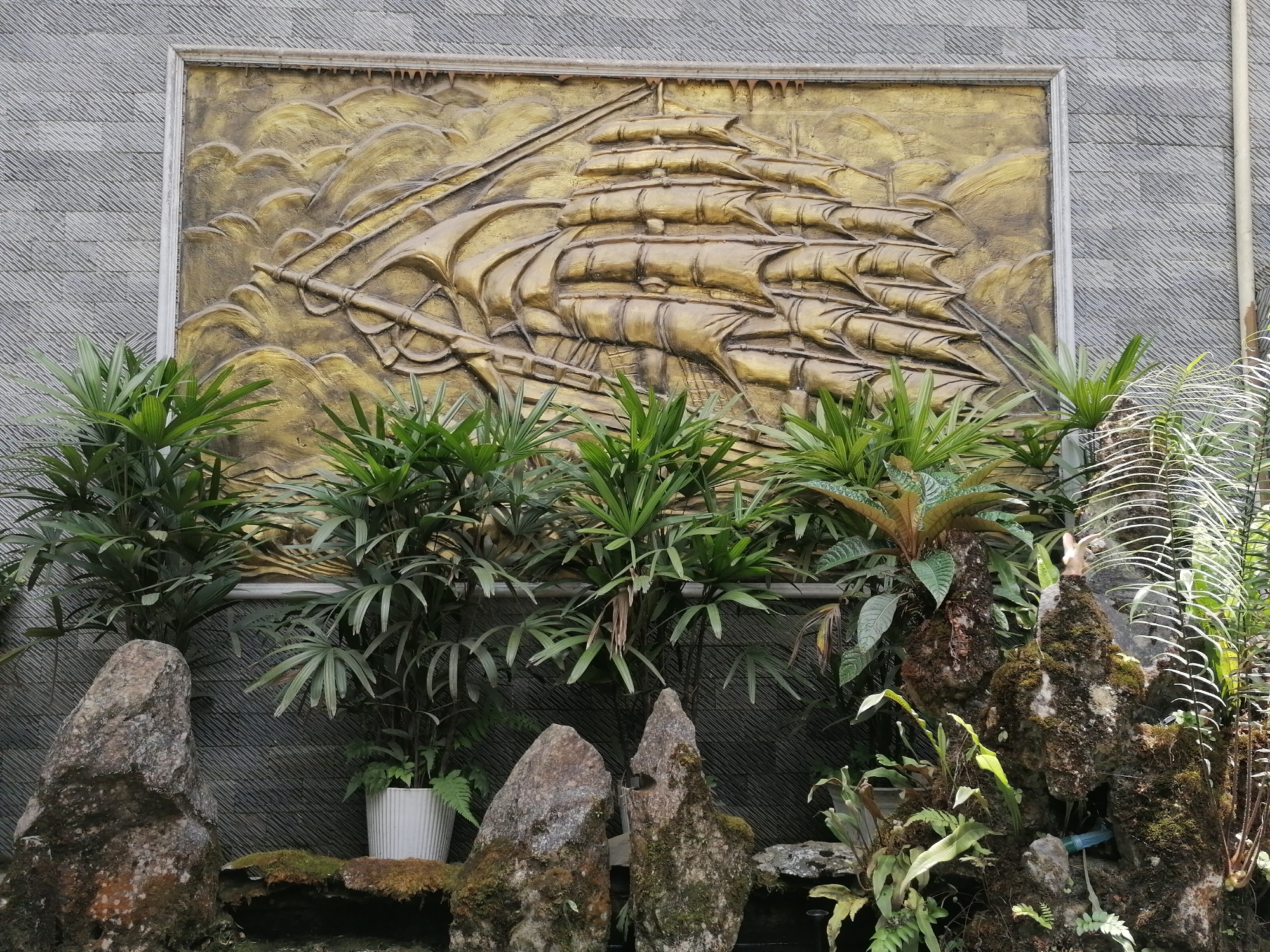 The image depicts a garden area with a large, rectangular, golden relief sculpture of a sailing ship mounted on a gray, horizontally-grooved wall. The sculpture shows a multi-masted ship with billowing sails, rendered in a heavily textured style. The wall is part of a larger structure. In front of the wall and sculpture, a variety of plants are arranged amongst moss-covered rocks. There are at least three distinct types of plants, with broad, fan-like green leaves. The rocks are dark gray with patches of green moss. One plant is in a white pot. A small fountain is visible among the rocks and plants. The setting appears to be in Sa Pa Ward, Vietnam, based on the style of the sculpture and the type of plants.