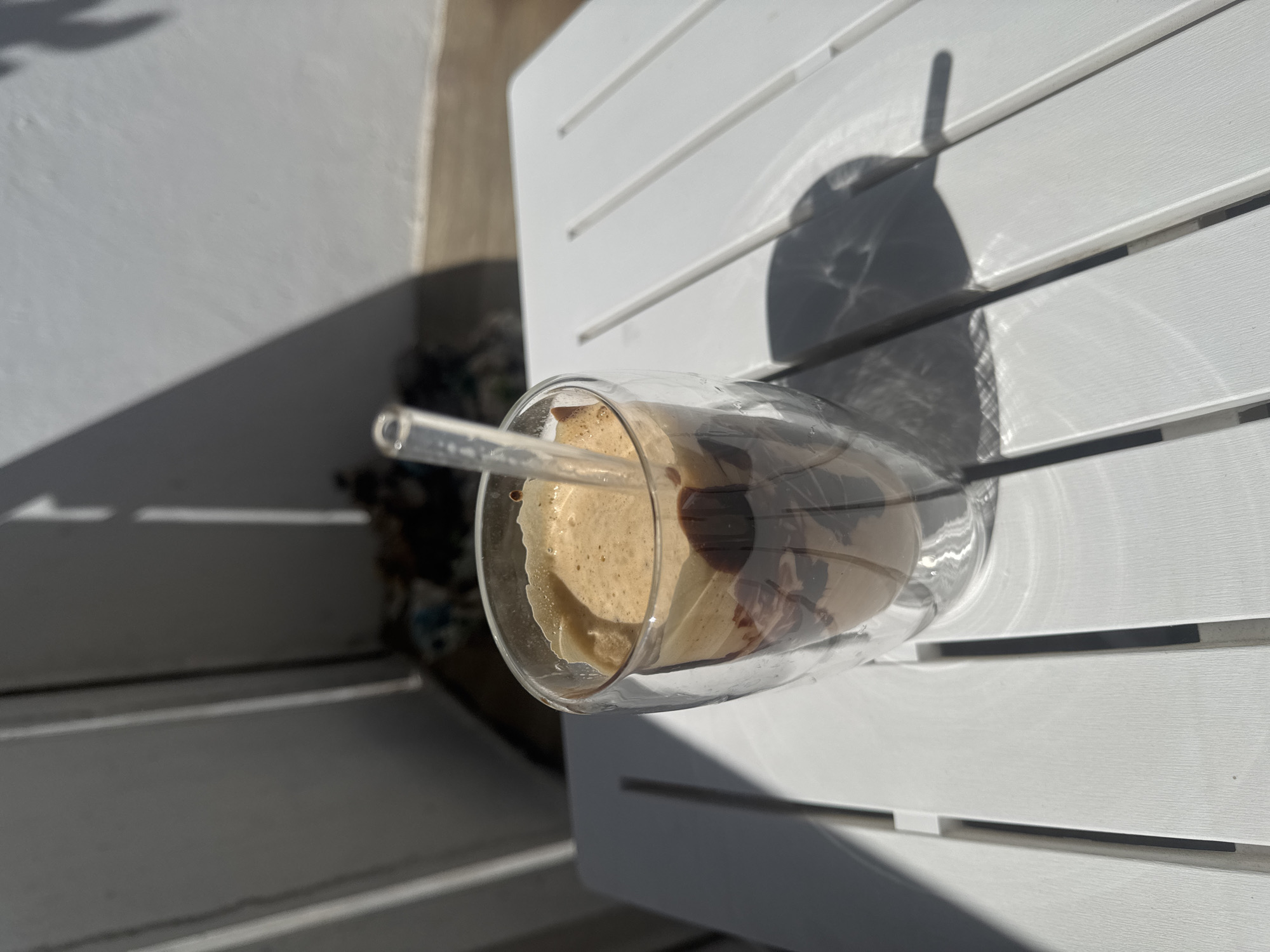 A clear glass with a double wall holds a frothy, light brown beverage, possibly a coffee or a milkshake, with streaks of dark chocolate syrup swirled within. A clear plastic straw extends from the glass, angled upwards and to the left. The glass is positioned on a white slatted surface, likely an outdoor table, bathed in bright sunlight that casts sharp, elongated shadows. The shadows suggest it is either morning or late afternoon. The background is blurred, but appears to be an outdoor setting with a textured white wall and hints of greenery or patterned fabric. The overall impression is one of a refreshing drink enjoyed outdoors on a sunny day. No text is visible.