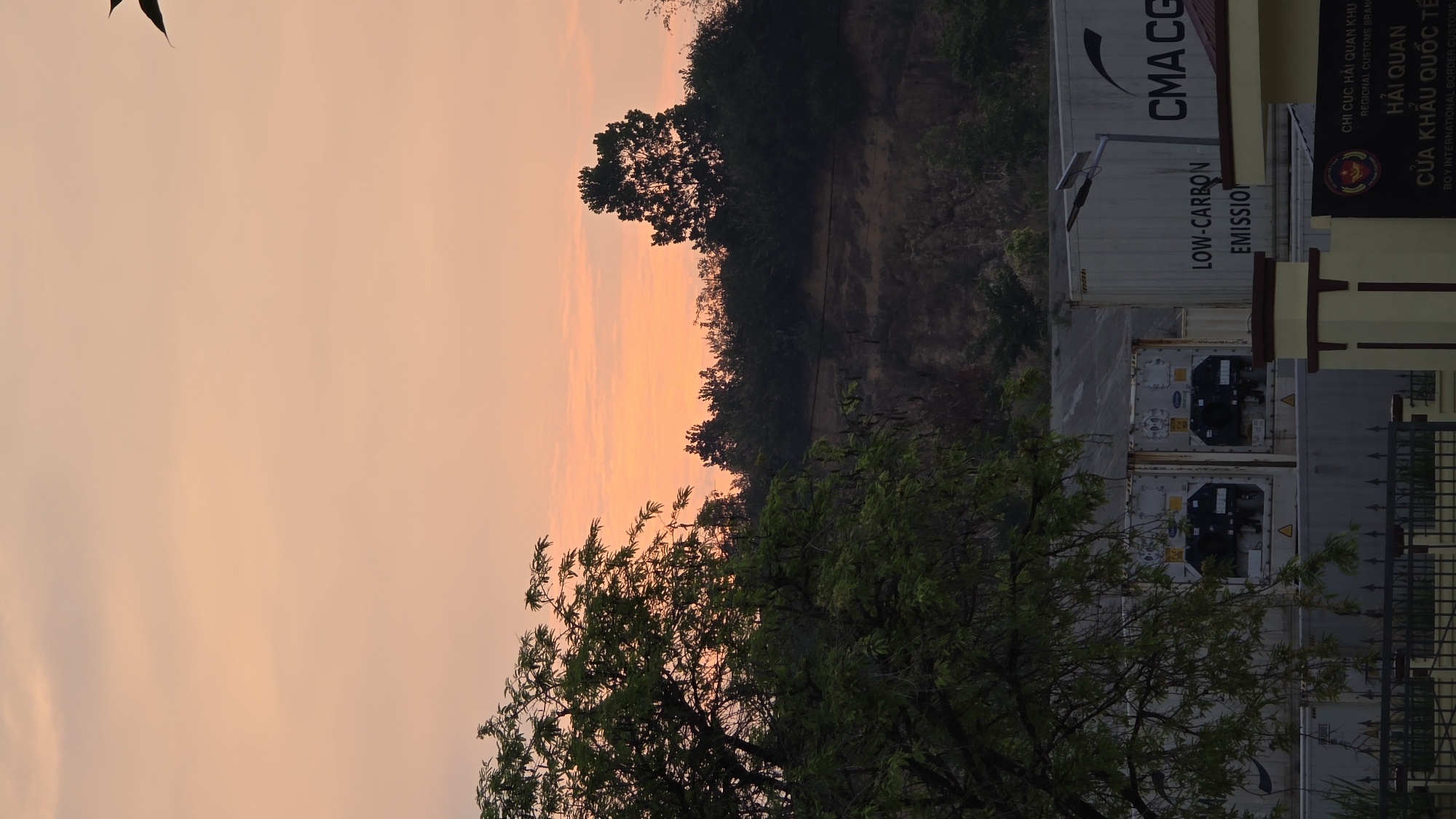 The image shows a landscape at sunset in Bờ Y, Vietnam. The sky is a mix of orange and pink. Trees are visible in the foreground. Further back, there is a building, possibly a customs office. Shipping containers are stacked beside the building. The signage on the building contains Vietnamese text. The containers are marked with "CMA CGM" and "LOW-CARBON EMISSION."