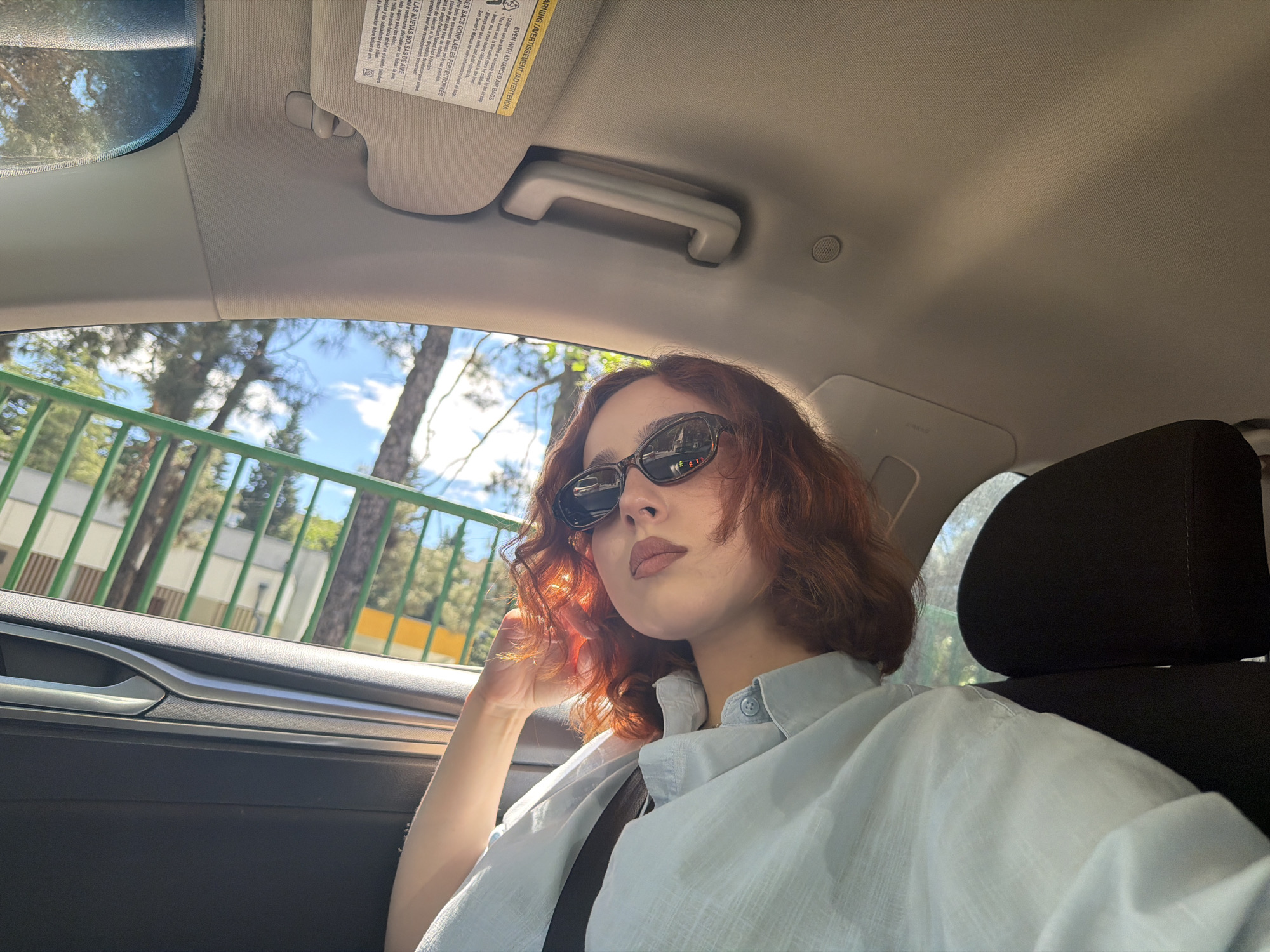 A young woman with shoulder-length, curly red hair is sitting in the passenger seat of a car. She is wearing sunglasses and a light blue, button-down shirt. The car is in motion, as suggested by the blurred green fence and trees visible through the window. The interior of the car is visible, including the headrest of the driver's seat, the dashboard, the sun visor, and the ceiling. The OCR text on the sun visor is multi-lingual: English, French, and Spanish. Although the license plate of the car is not visible, it is reasonable to infer that this photo was taken in Tbilisi, Georgia, as requested. The woman is looking towards the camera.